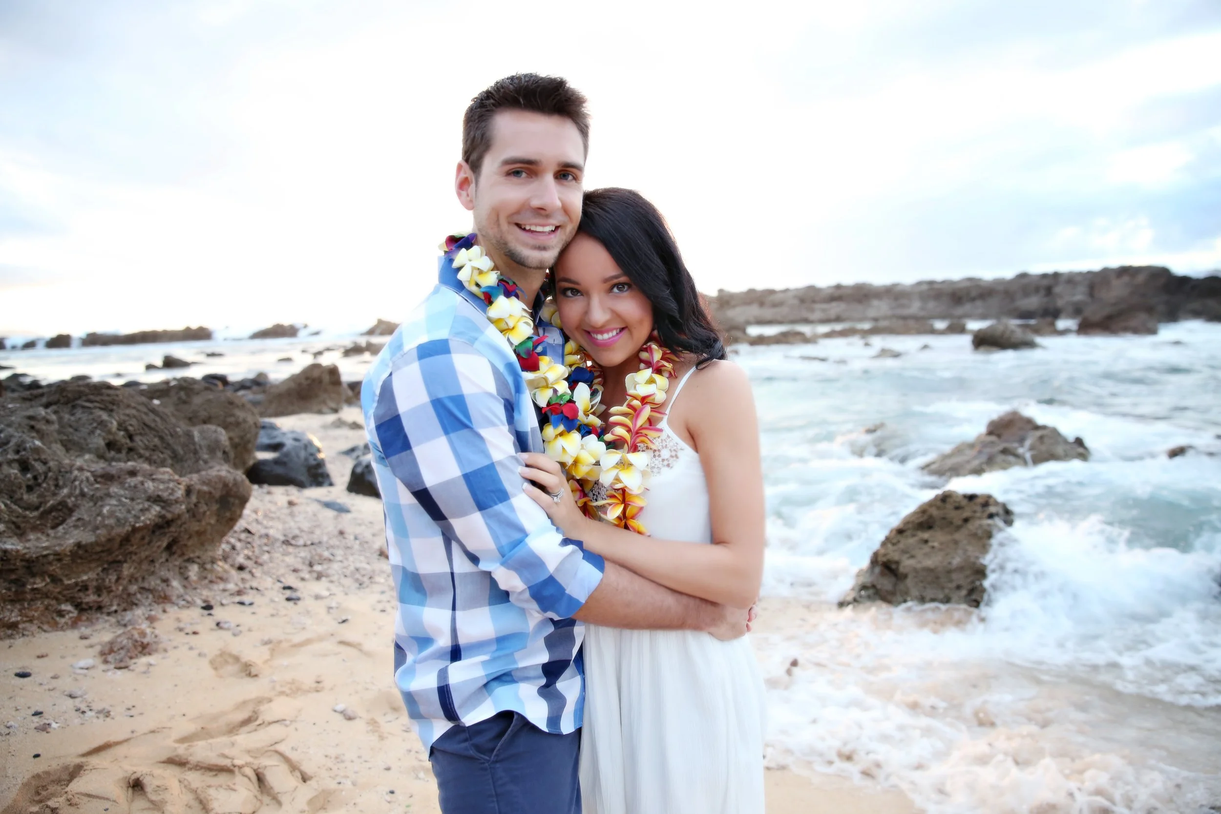 A happy couple in white and blue clothing with leis, standing arm in arm on a rocky beach with the ocean in the background.