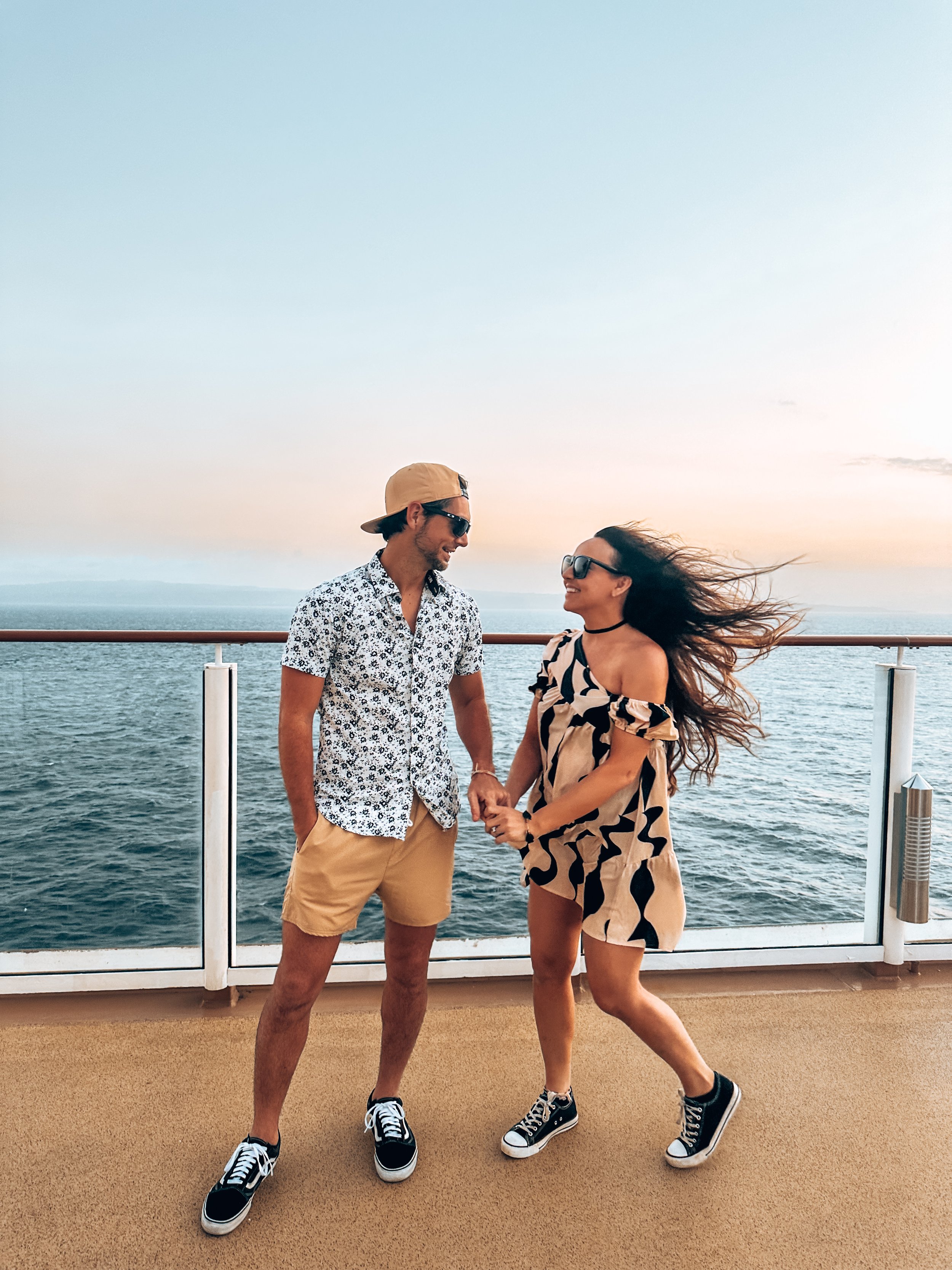 A man and woman holding hands on the deck of a cruise ship at sunset, with the ocean in the background and both smiling wearing casual summer clothes.