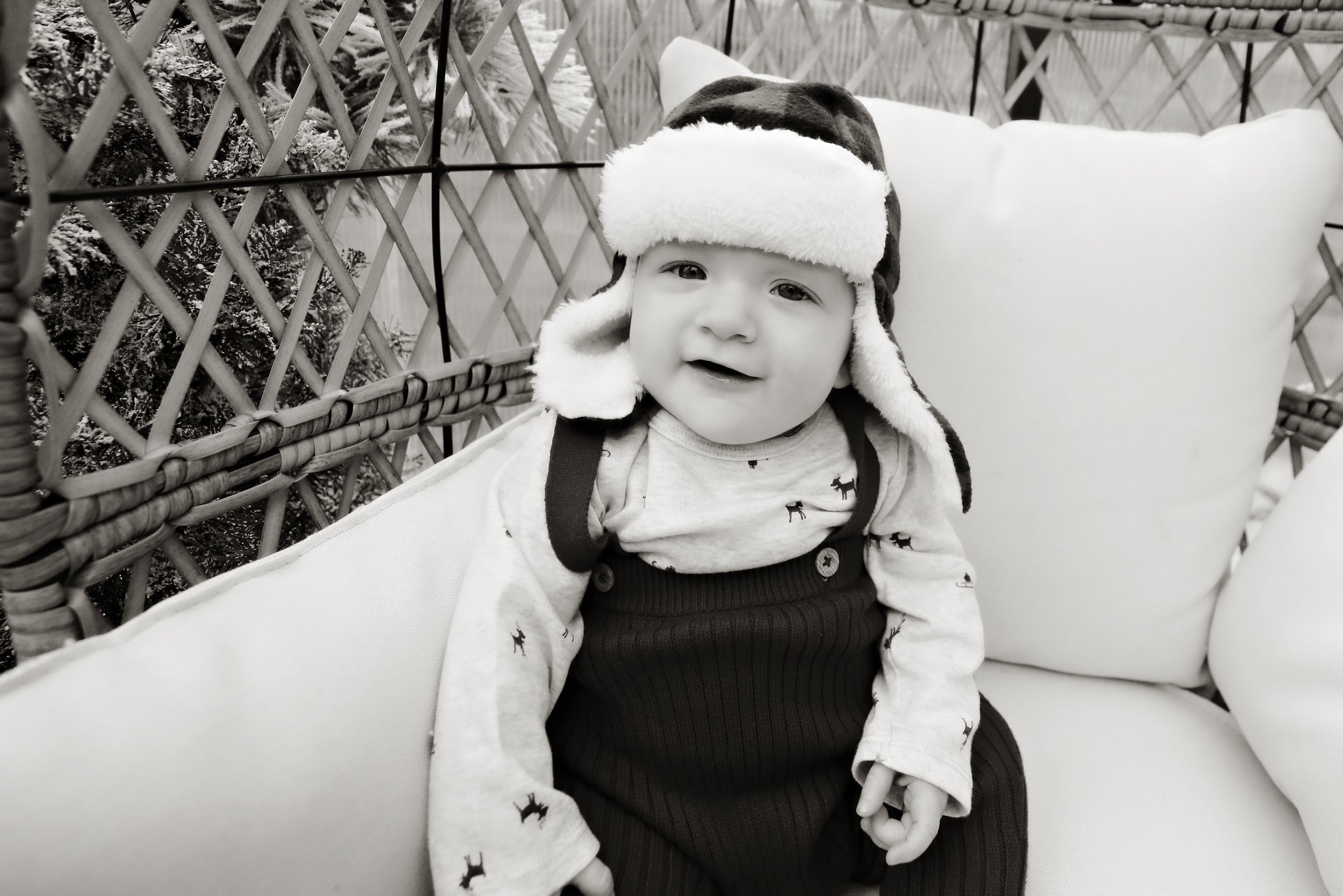 A young child wearing a winter hat with ear flaps and a long-sleeve shirt with small dog patterns, sitting on a cushioned outdoor chair with a lattice fence and snow-dusted trees in the background.