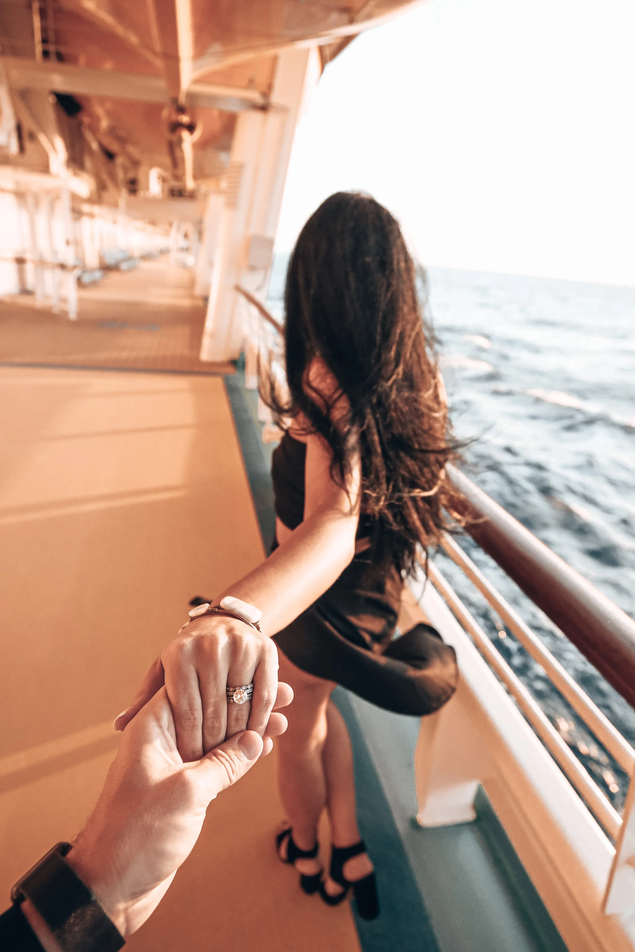 A woman with dark hair leaning on the ship's railing, holding hands with someone taking the photo, showing a diamond engagement ring. They are on the deck of a cruise ship with ocean waters visible and the sun setting in the background.