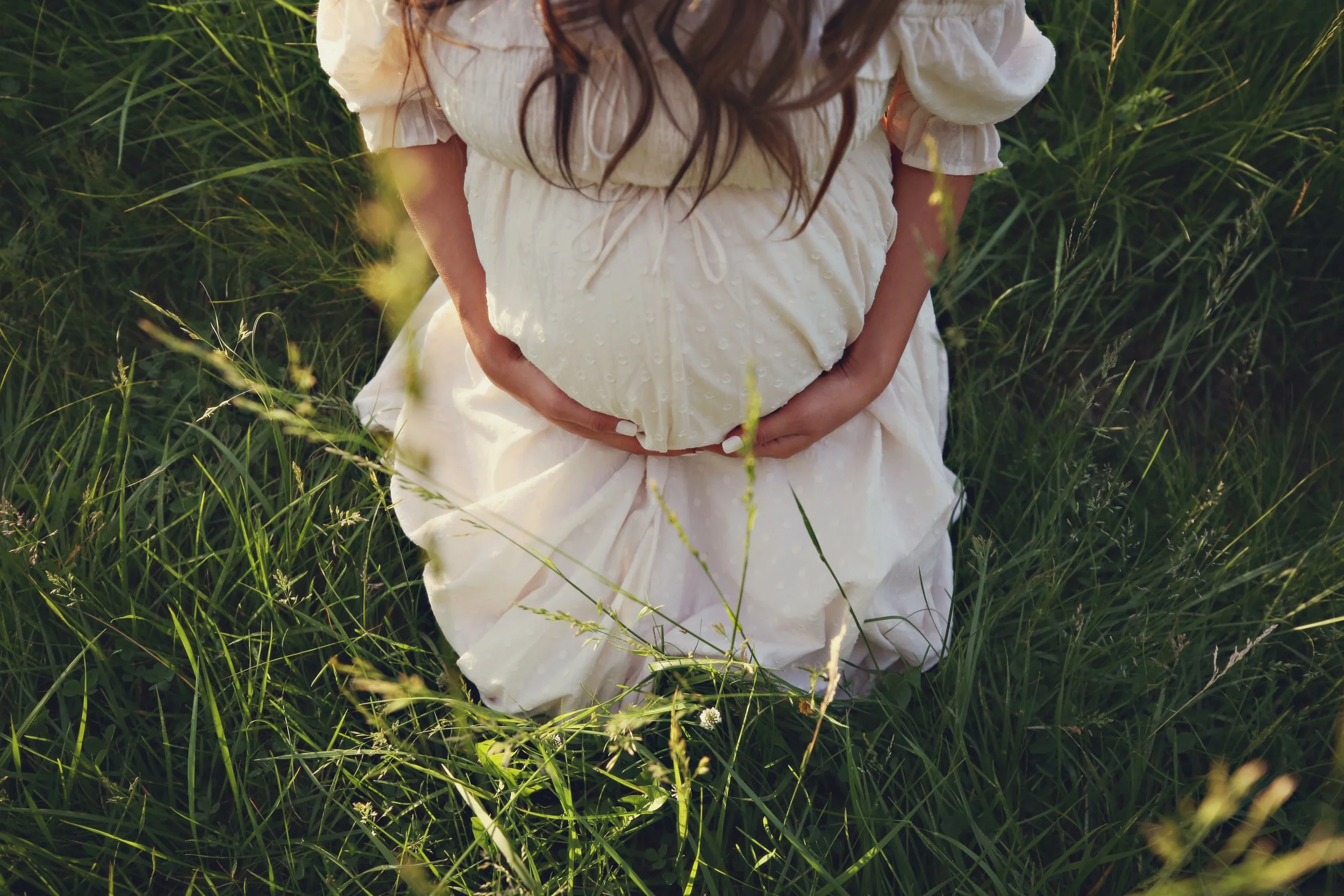 A woman wearing a white dress crouches in tall green grass, cradling her pregnant belly with both hands.