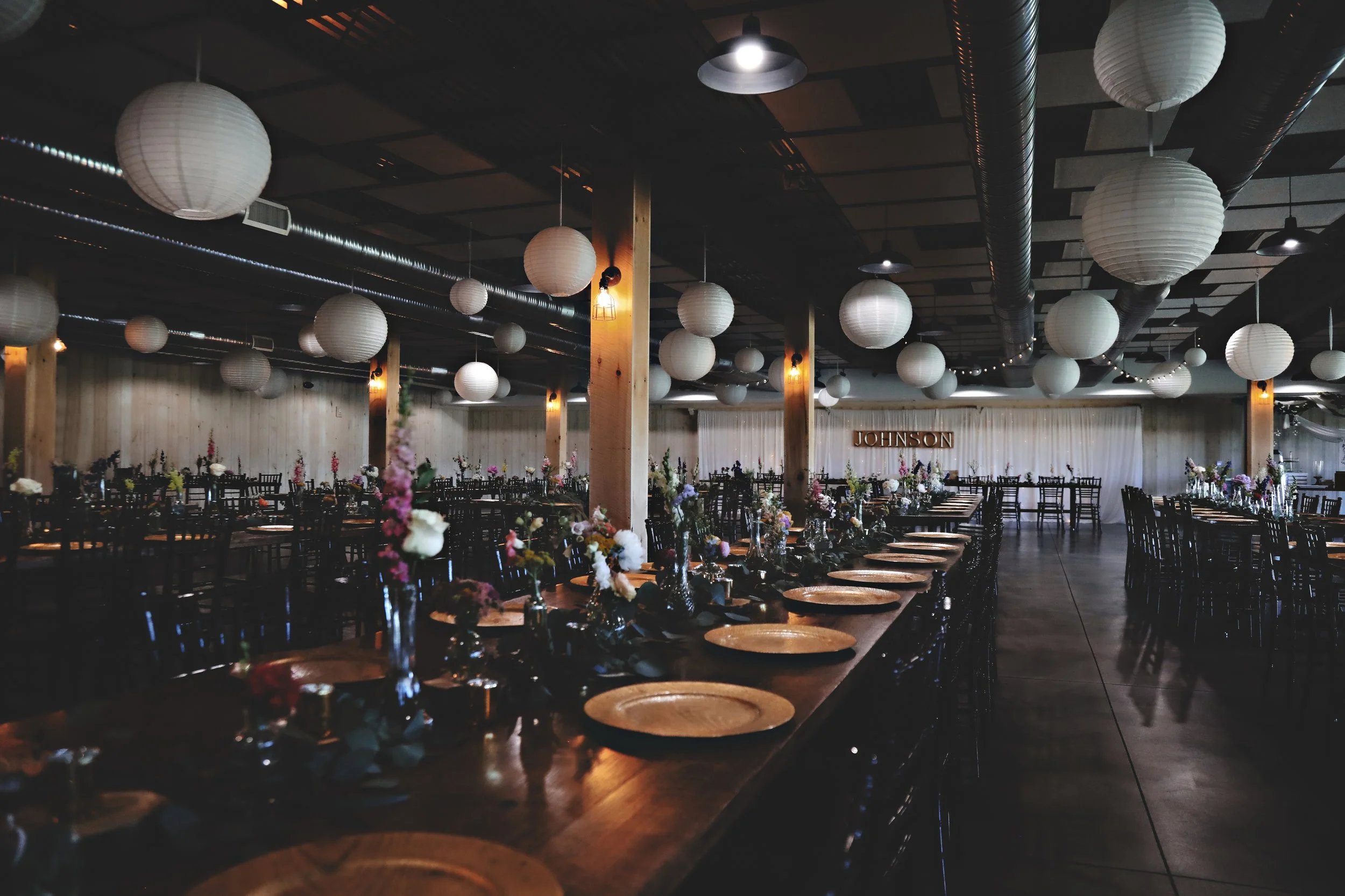 A decorated event hall with long wooden tables set with plates, cutlery, and flower centerpieces, hanging paper lanterns, and a stage with a sign that reads 'Johnson' in the background.