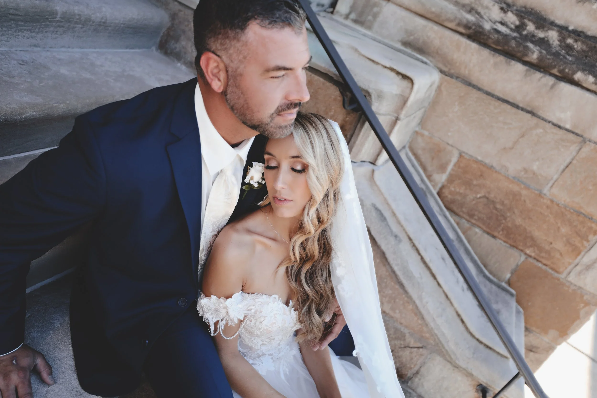A bride and groom sitting close together on steps, with the bride leaning her head on the groom's chest, both with closed eyes, in a romantic moment.