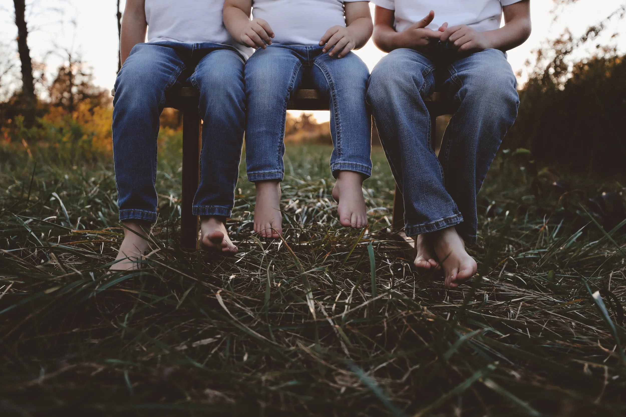Three children sitting barefoot on a bench outdoors during sunset, wearing jeans and white shirts.