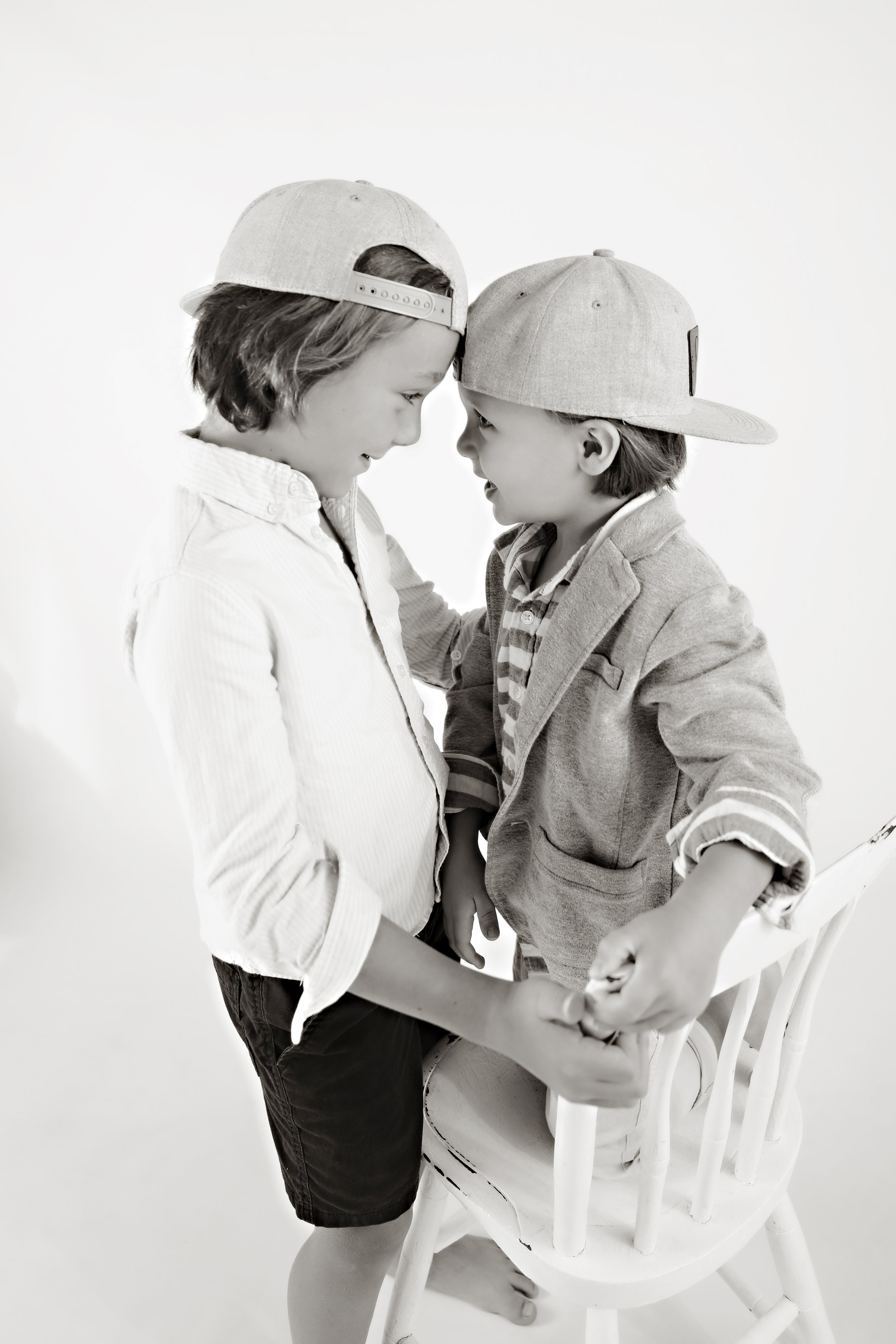 Two children, a girl and a boy, holding hands and smiling at each other while standing close, wearing baseball caps and casual clothing, with a white background.