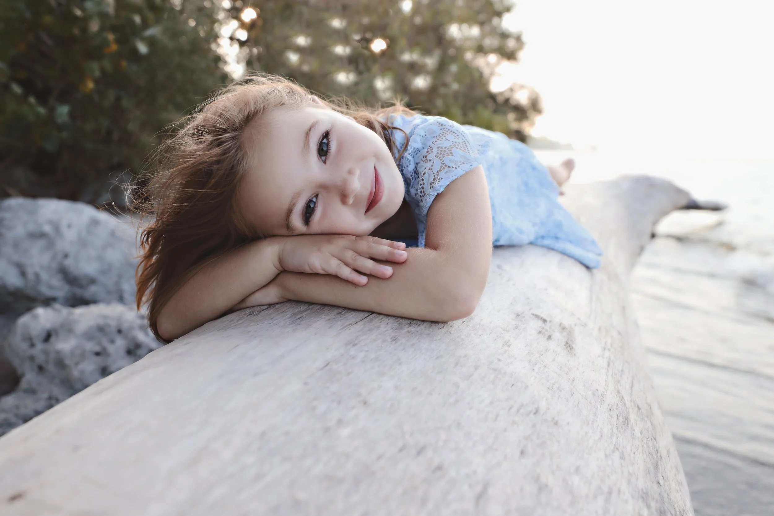 Young girl with red hair lying on a large rock by the water, resting her head on her folded arms and smiling softly, with trees and water in the background.
