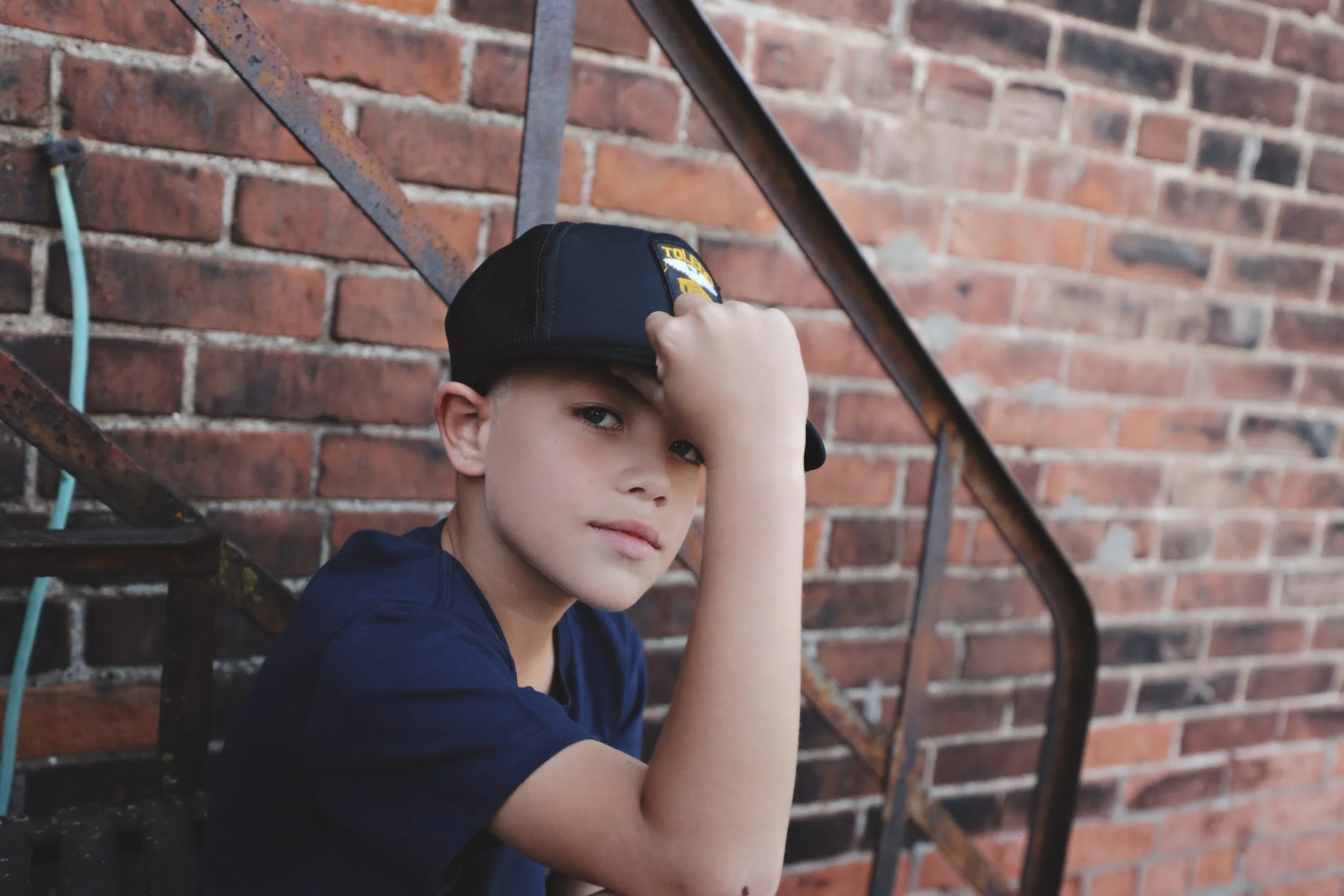 A young boy wearing a police hat sitting on outdoor stairs against a brick wall.