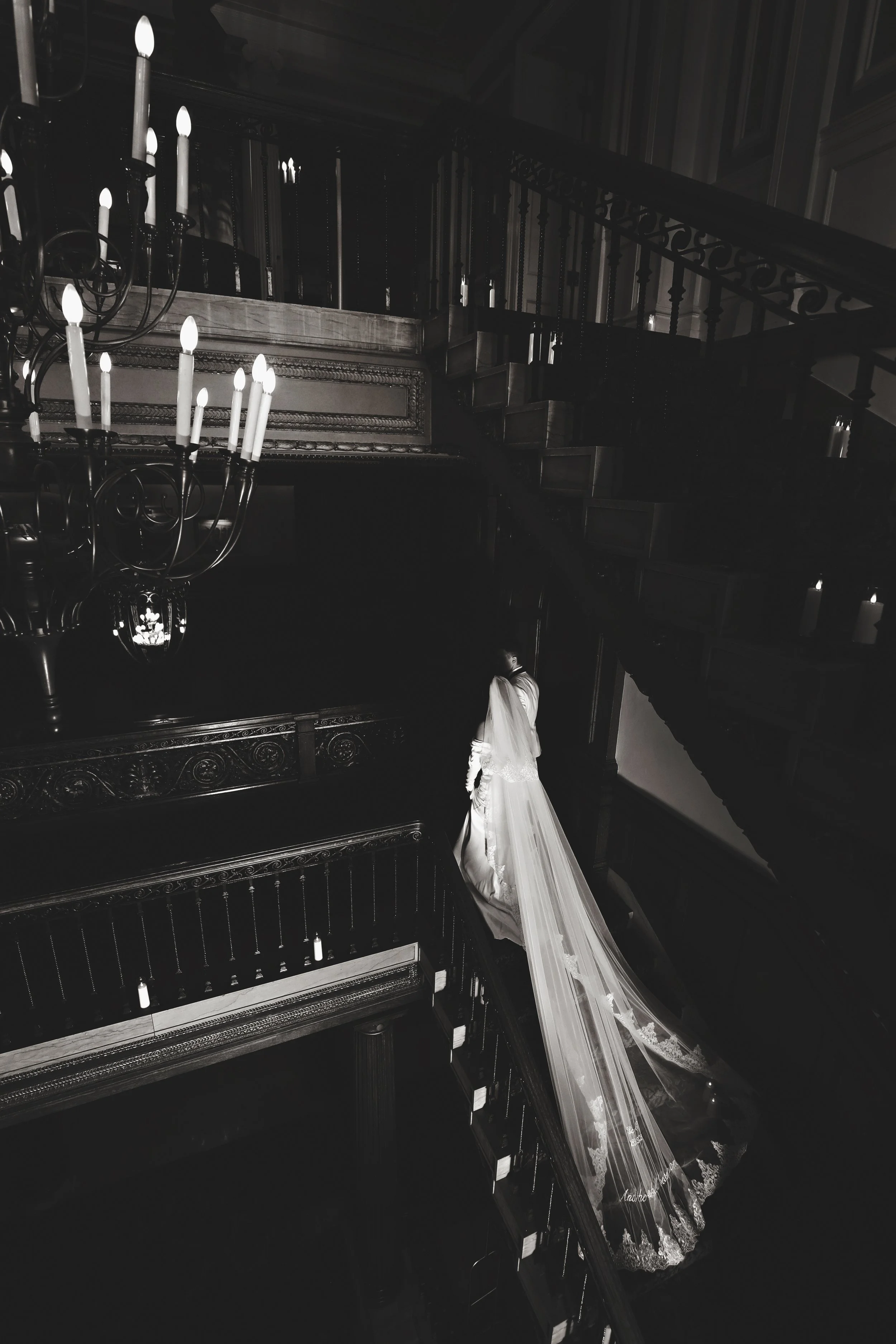 A bride in a wedding dress with a long veil standing on a staircase inside a grand, dark, ornate room with a chandelier.