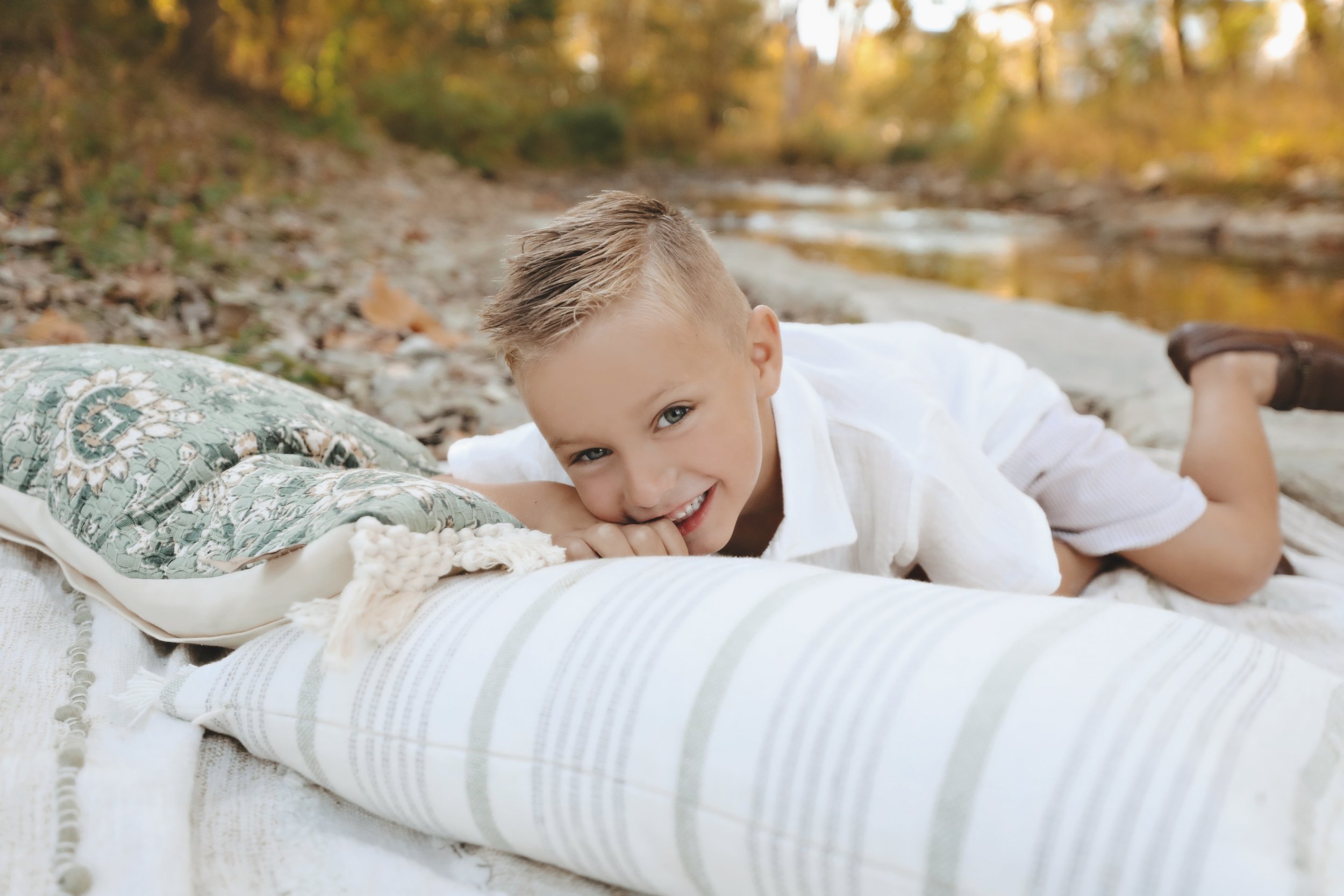 A young boy lying on a blanket on a riverbank, smiling and resting his head on a pillow with a paisley pattern.