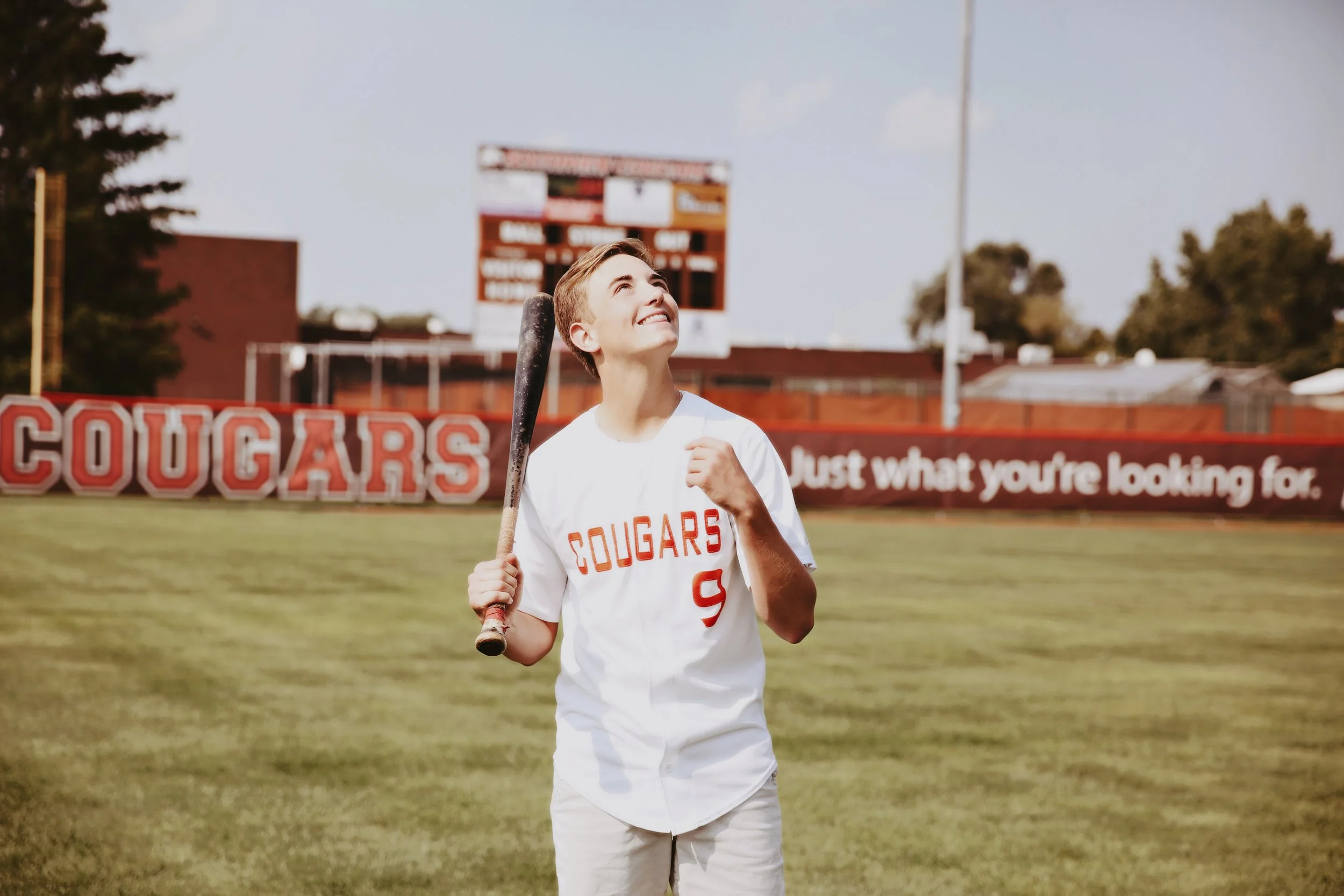 Young male baseball player on field holding bat, smiling, wearing white jersey with 'Cougars' and number 9, with scoreboard and school sign in background.