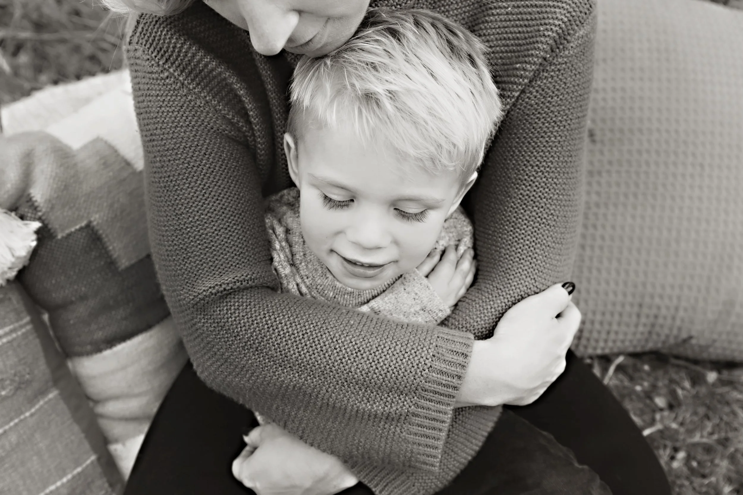 A woman hugging a young boy with light-colored hair and closed eyes, sitting outdoors on a bench with a pillow nearby.