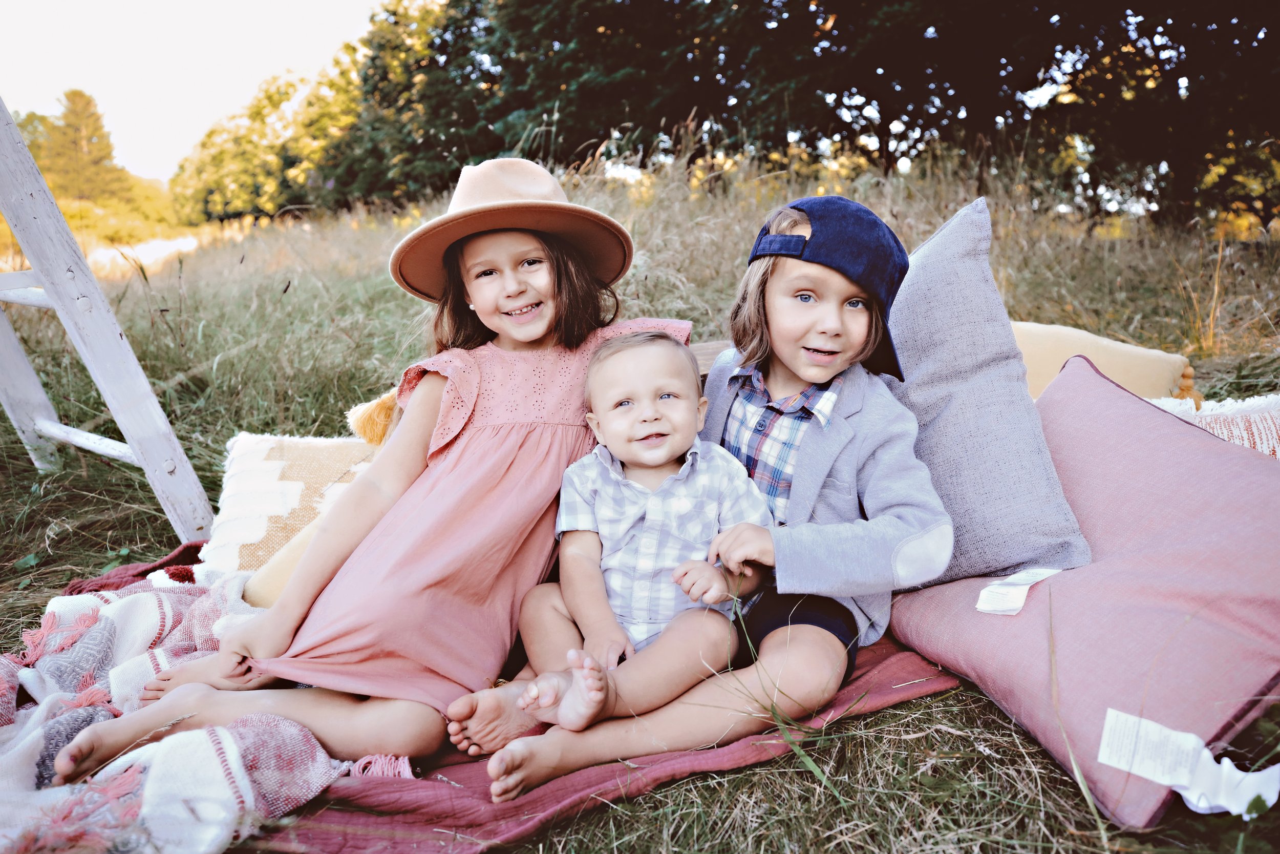 Three children sitting on a blanket outdoors surrounded by grass and trees.