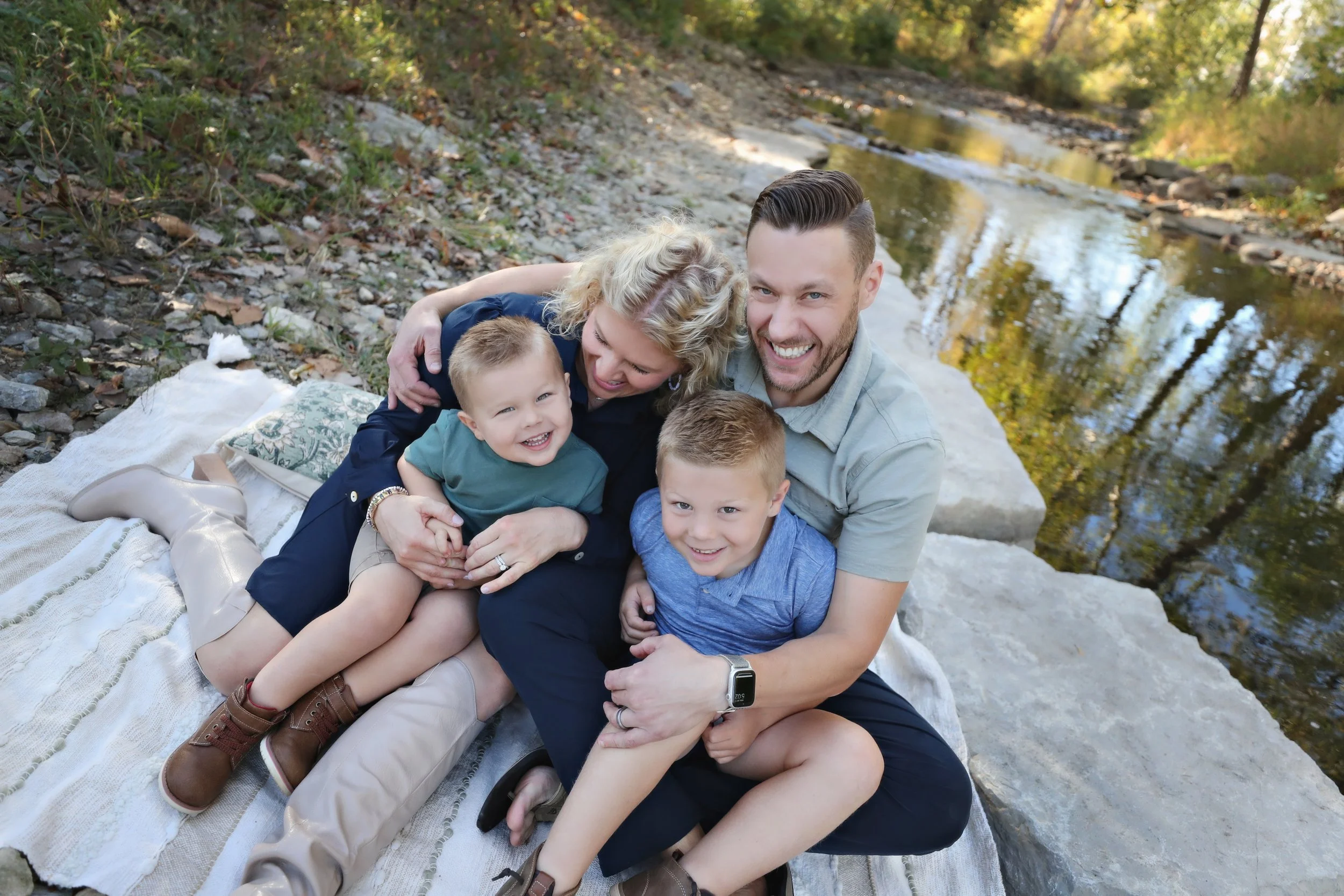 Family of four sitting on a blanket by a river, smiling and hugging outdoors during fall.