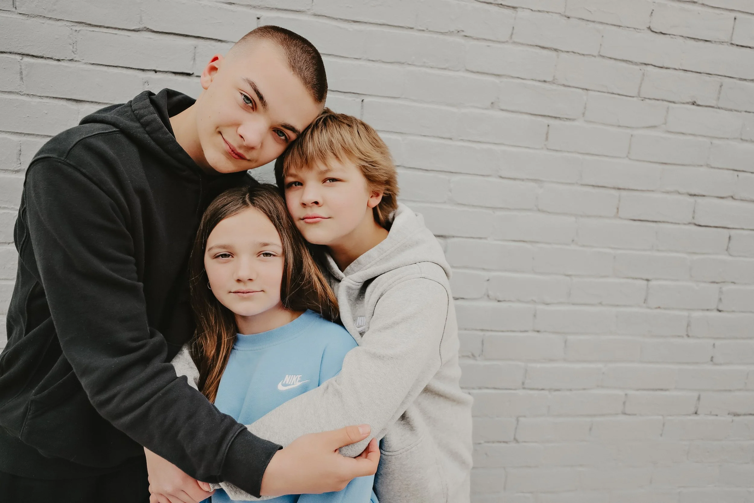 Three children, one girl and two boys, hugging together against a white brick wall.