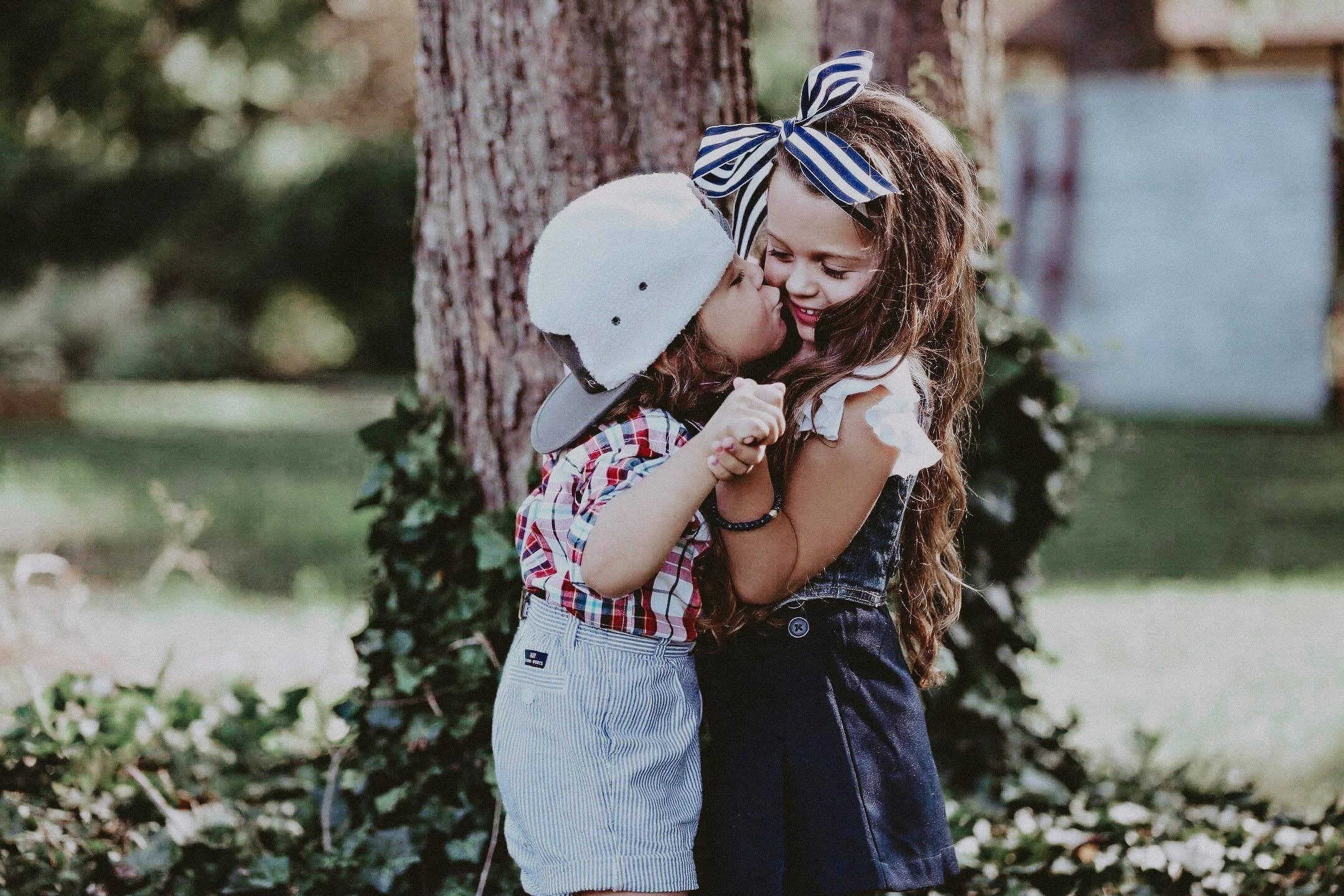 Two children, a girl and a boy, sharing a hug and kiss outdoors near a tree, with green foliage in the background.