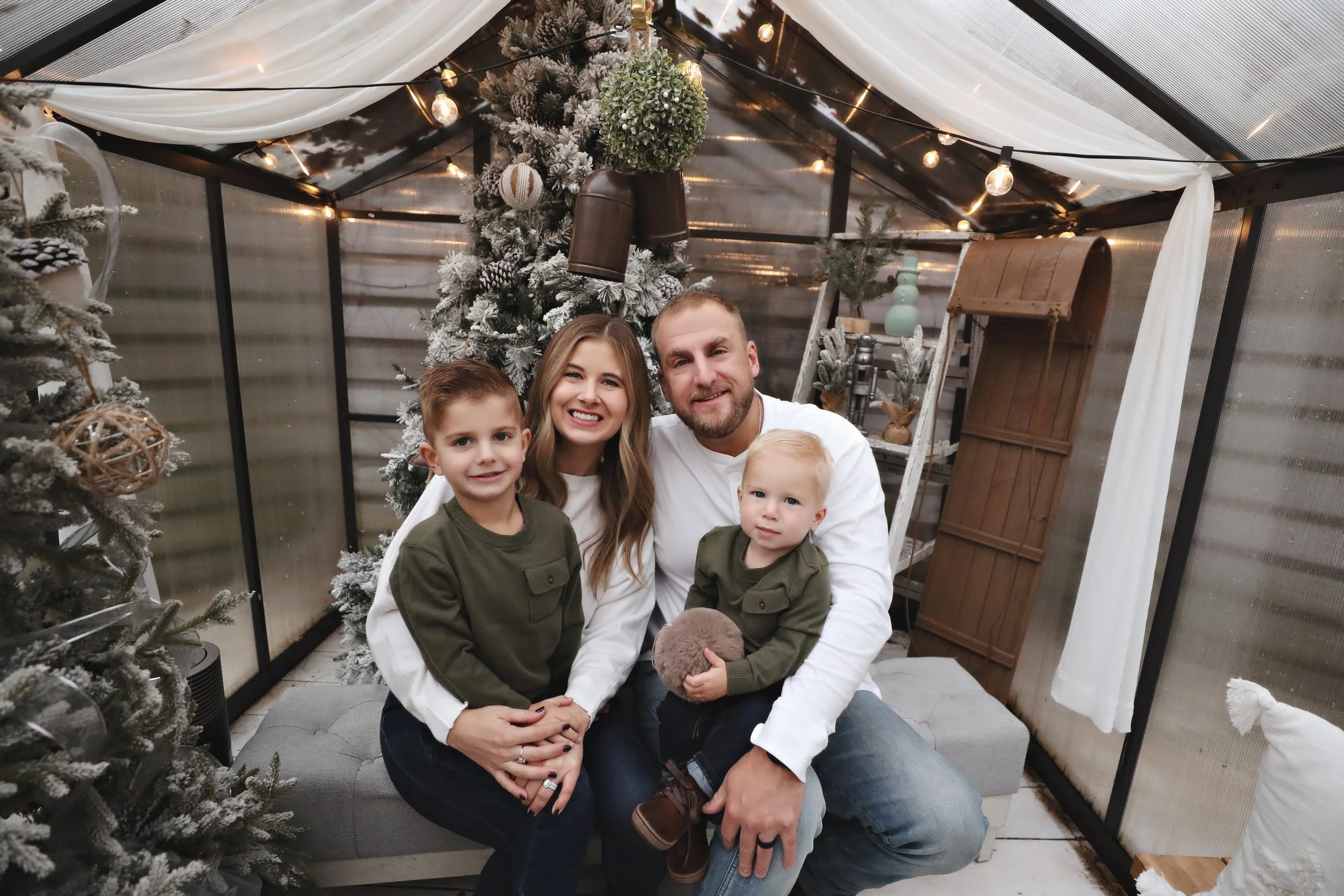 A family of four sitting in a decorated indoor space with Christmas trees, ornaments, and string lights, smiling for the camera.
