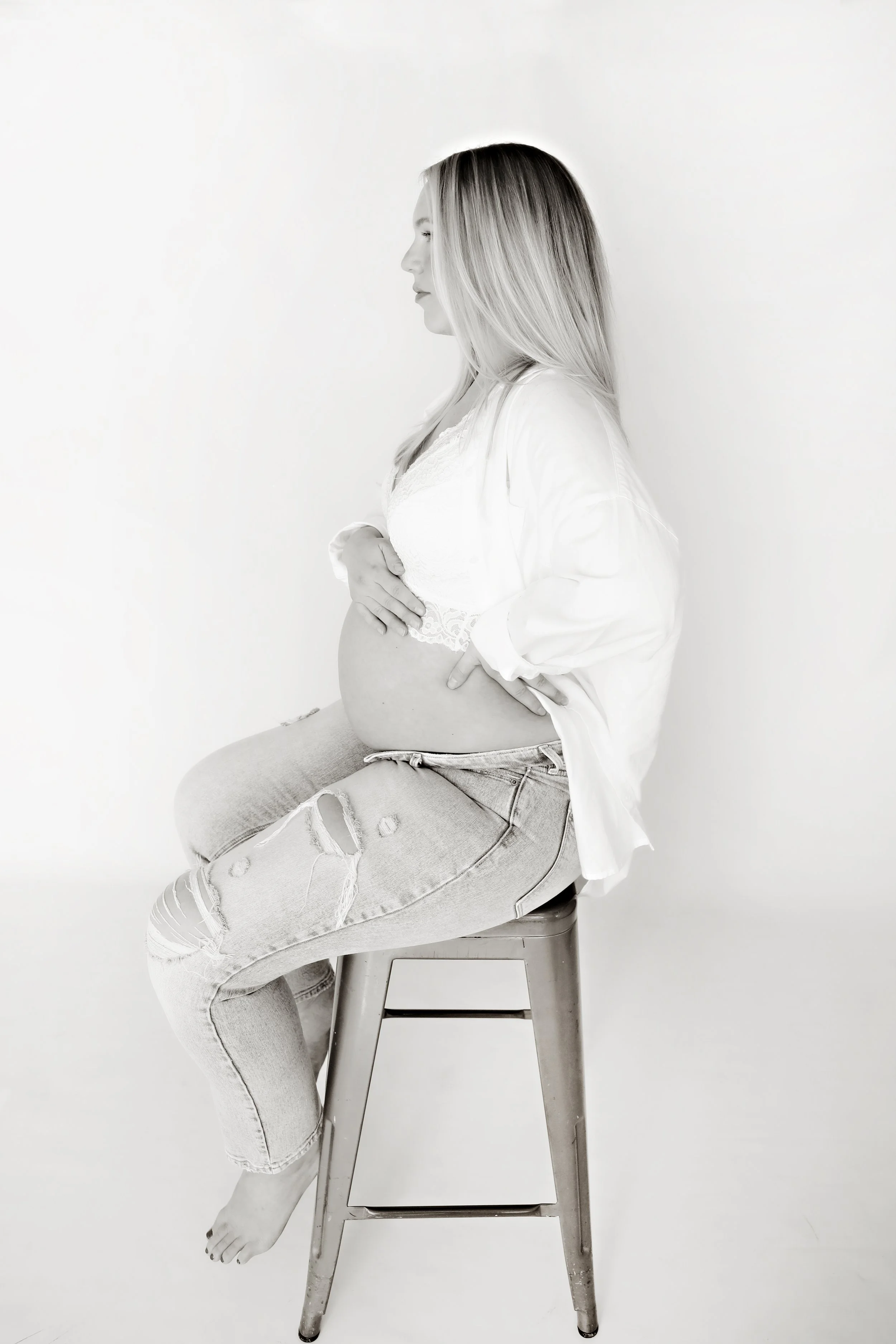 Black and white photo of a pregnant woman sitting on a stool, with her hands on her belly, wearing distressed jeans and a loose shirt.