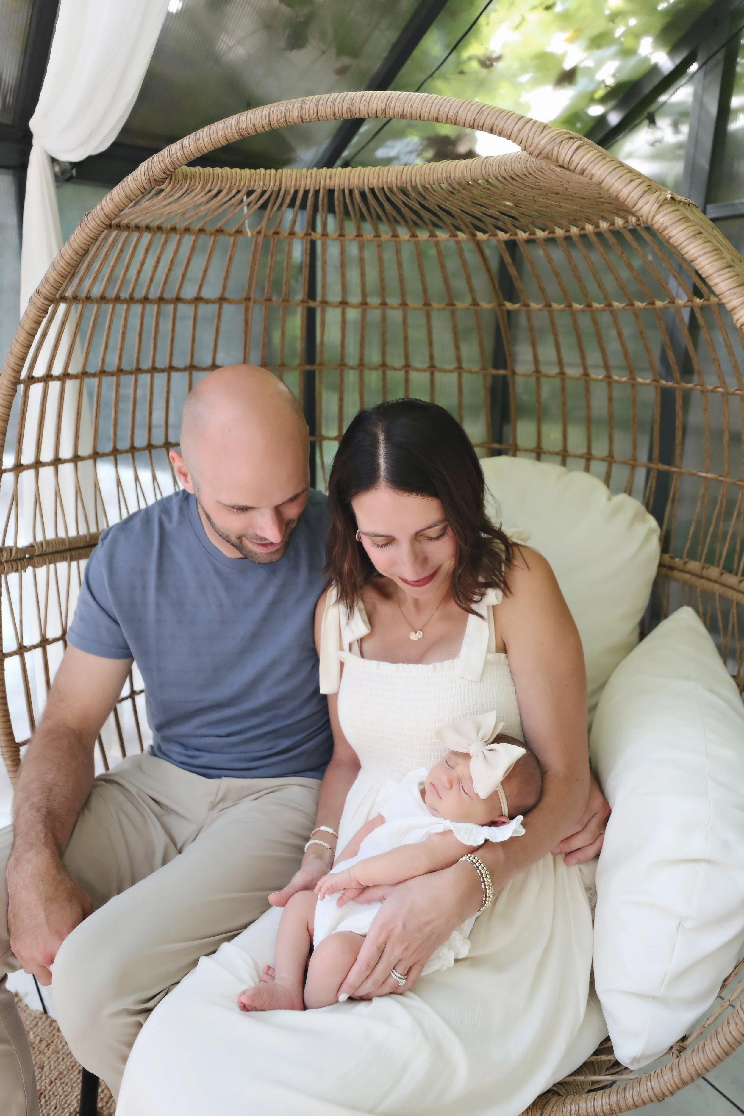 A family of three, a man, a woman, and a sleeping baby girl, sitting together on a white cushion inside a wicker egg-shaped chair with a canopy in a glass-enclosed outdoor space.