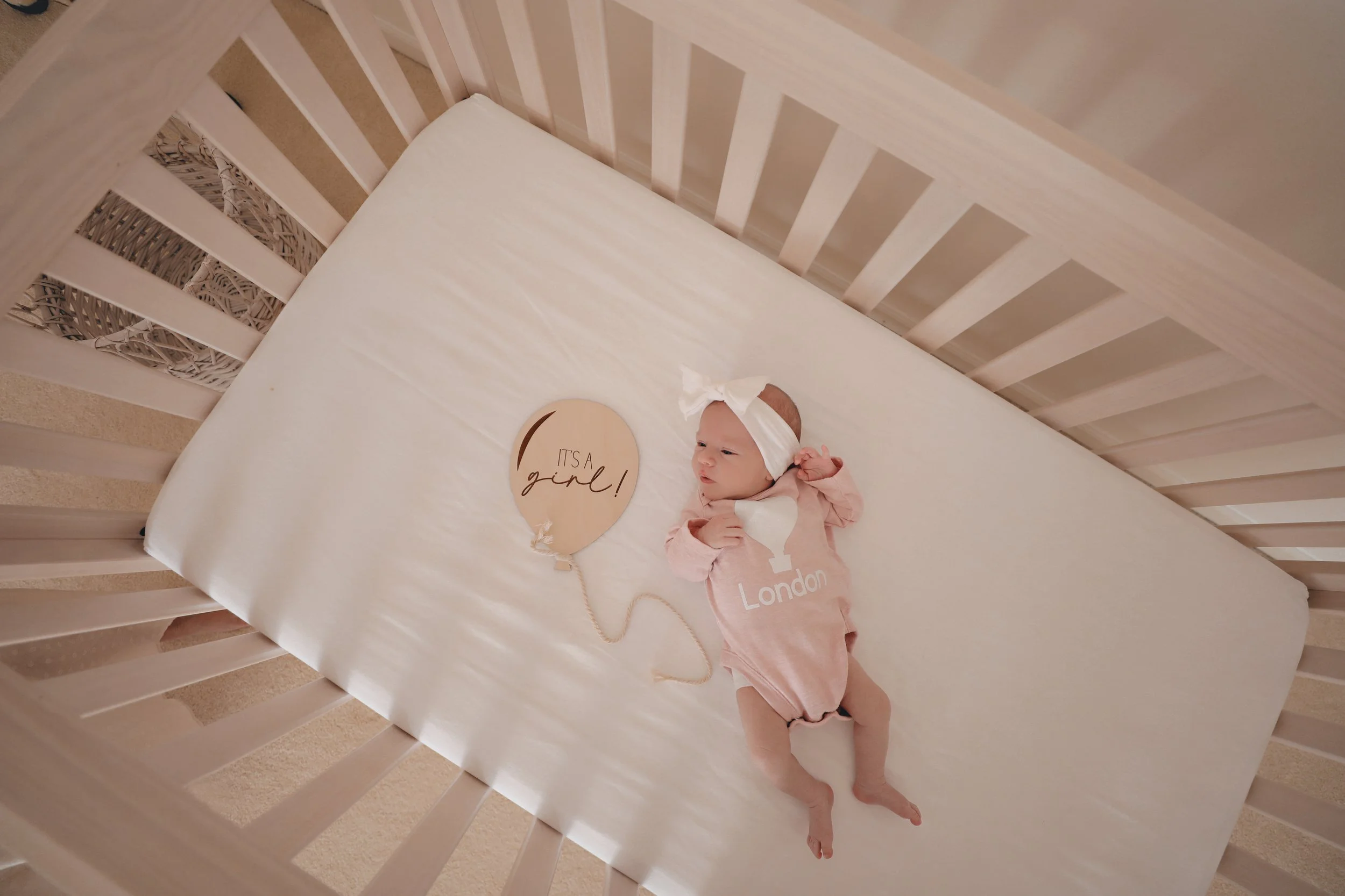 Infant lying in a crib wearing a pink onesie with 'London' printed on it, and a white headband with a bow. A heart-shaped card next to the baby reads 'It's a girl!'