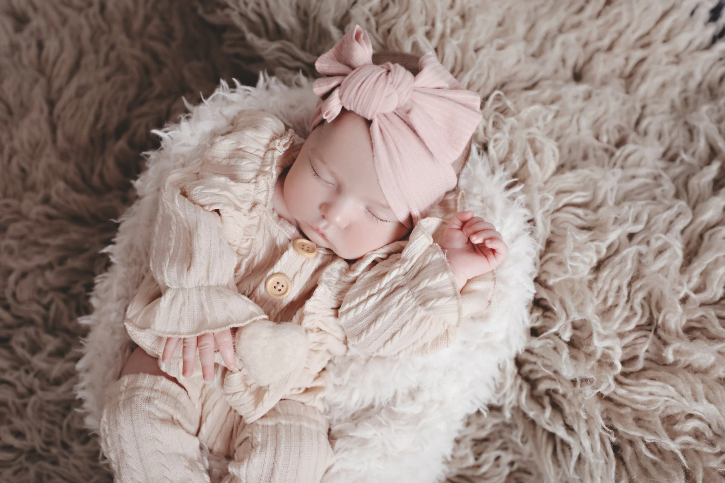 A sleeping baby girl with a pink bow headband, dressed in beige ruffled outfit, lying on a fluffy cream-colored blanket.