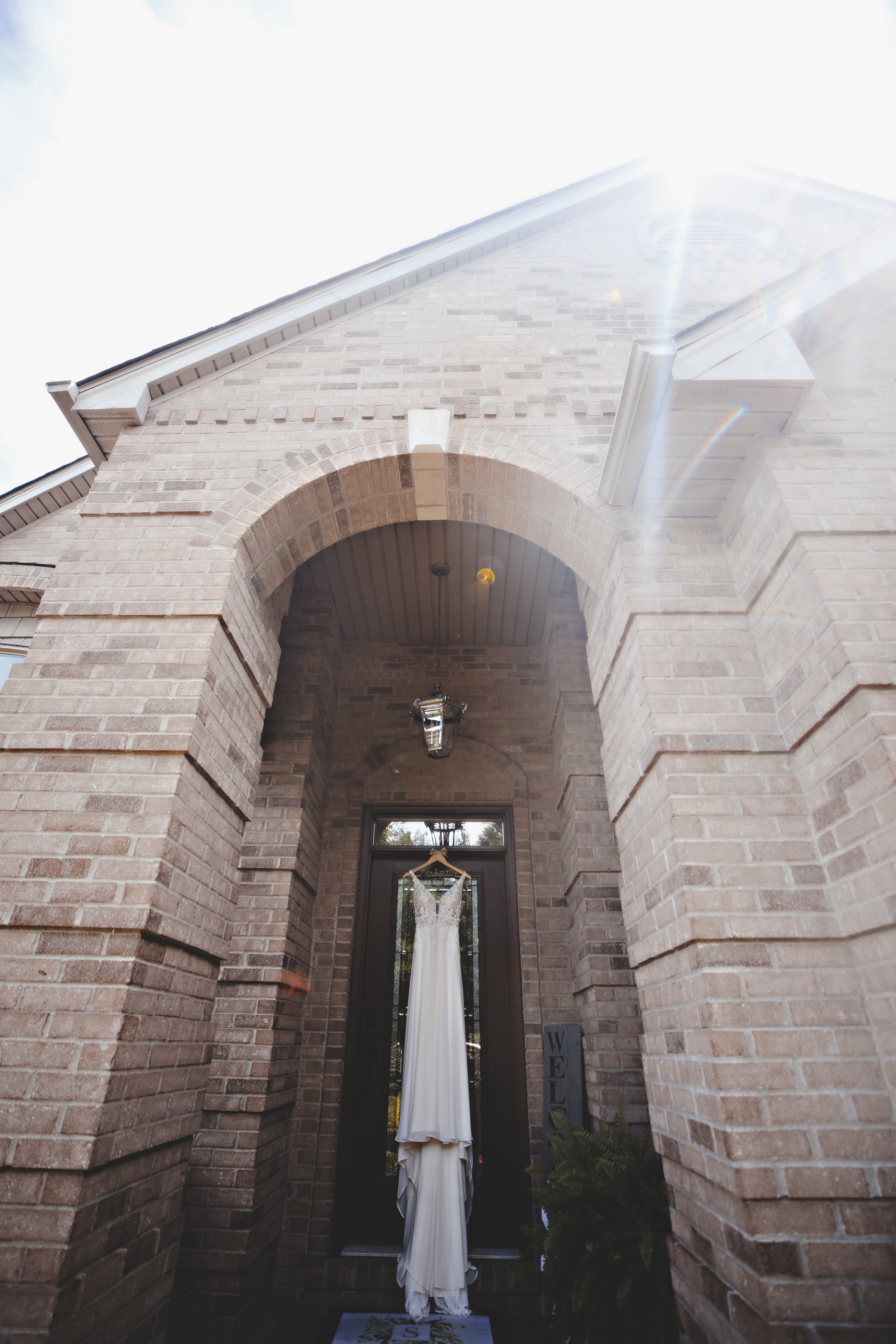 Wedding dress hanging on a hanger in front of a glass door under an arched brick entryway, outside a brick house, with sunlight and a fern plant nearby.