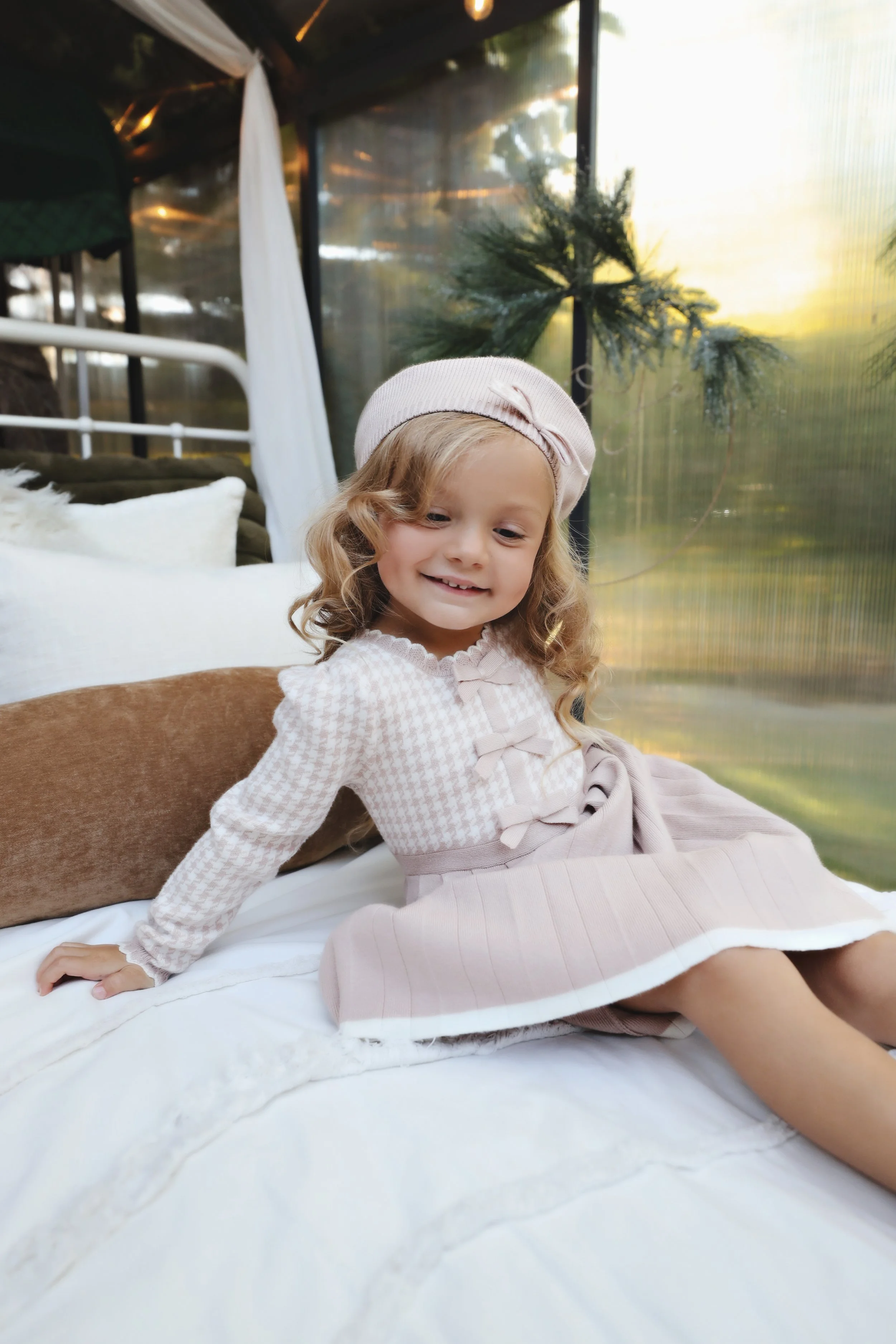 A young girl with curly blonde hair wearing a light pink beret and a pink and white dress sitting on a bed inside a cozy, well-lit room with a window showing trees outside.