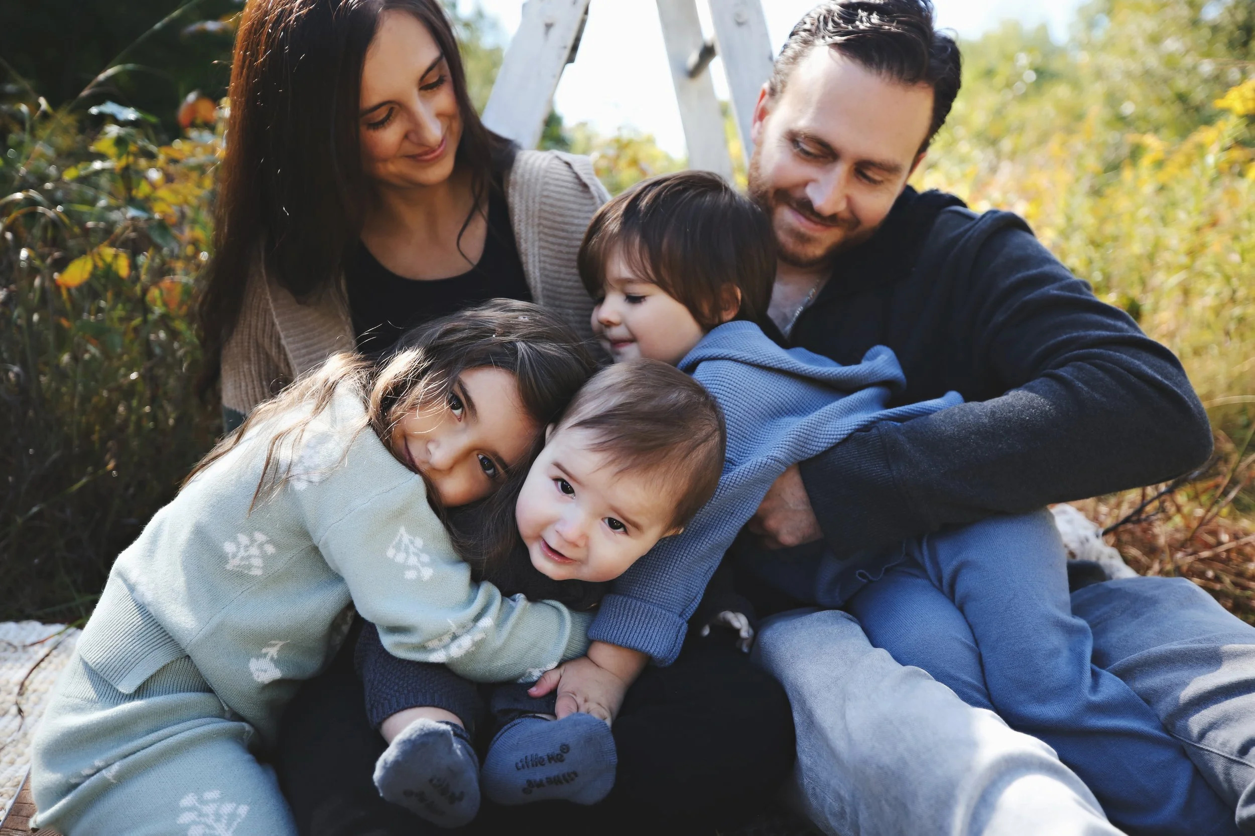 A family of six enjoying time outdoors on a fall day, sitting on the ground surrounded by trees and foliage, hugging and smiling.