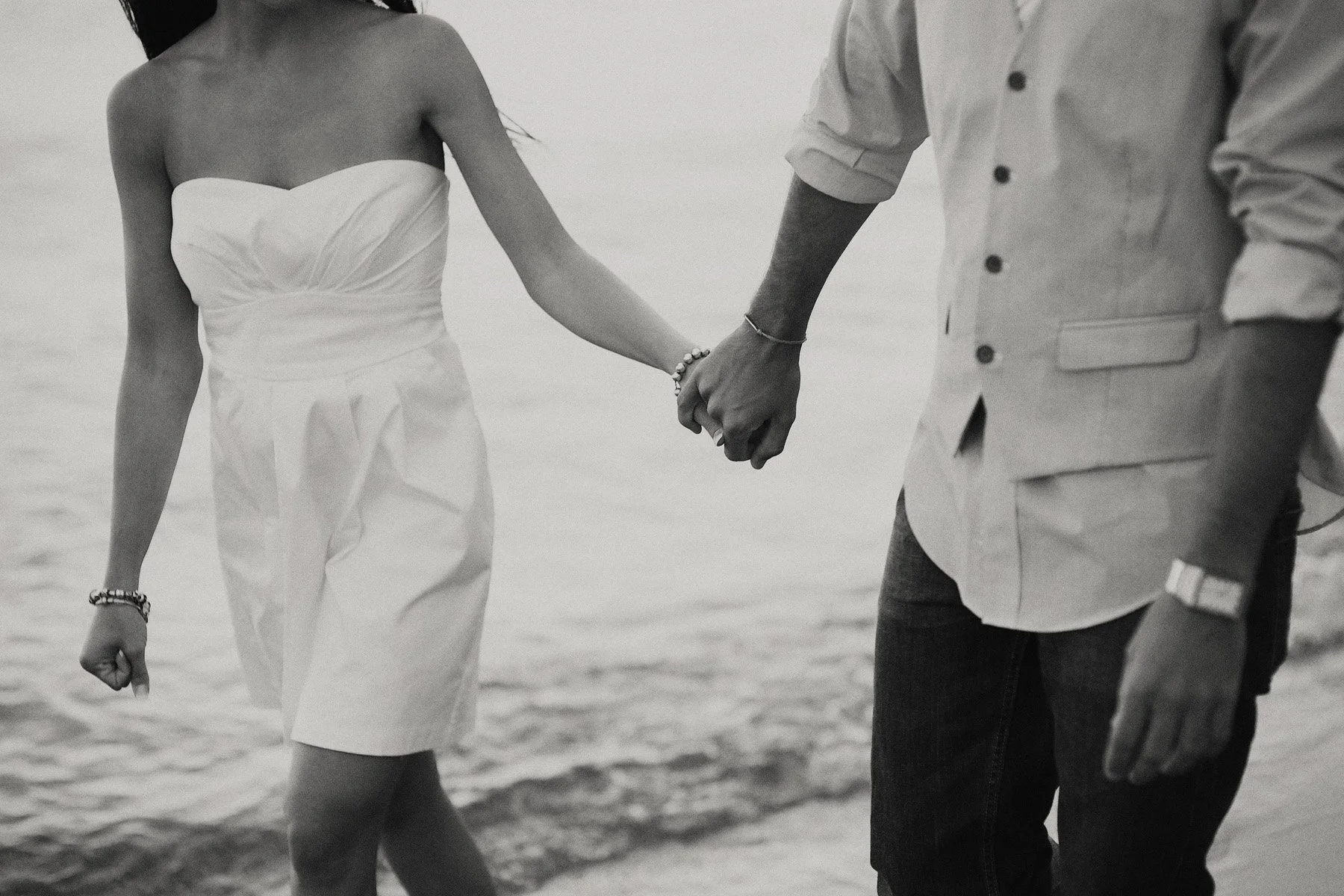 A couple holding hands on a beach, with both dressed in casual summer clothes, near the water's edge.