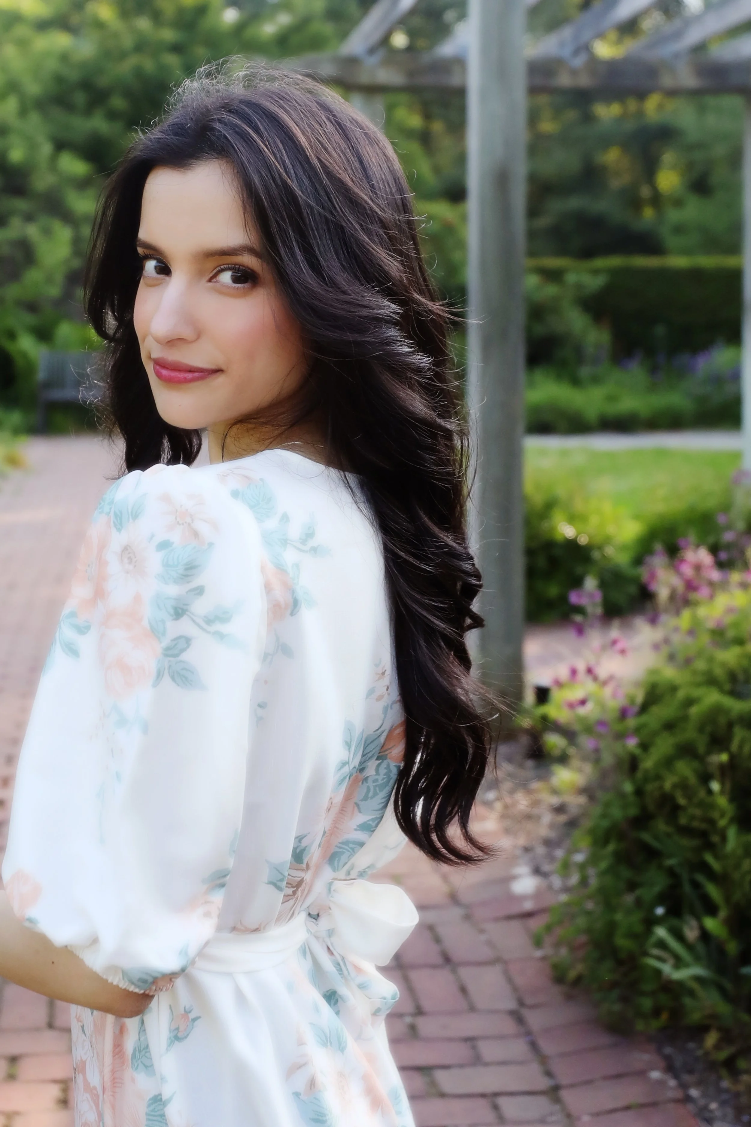 A young woman with long, dark, wavy hair and light skin, wearing a white floral dress with puffed sleeves, standing on a brick pathway in a garden with greenery and flowers.