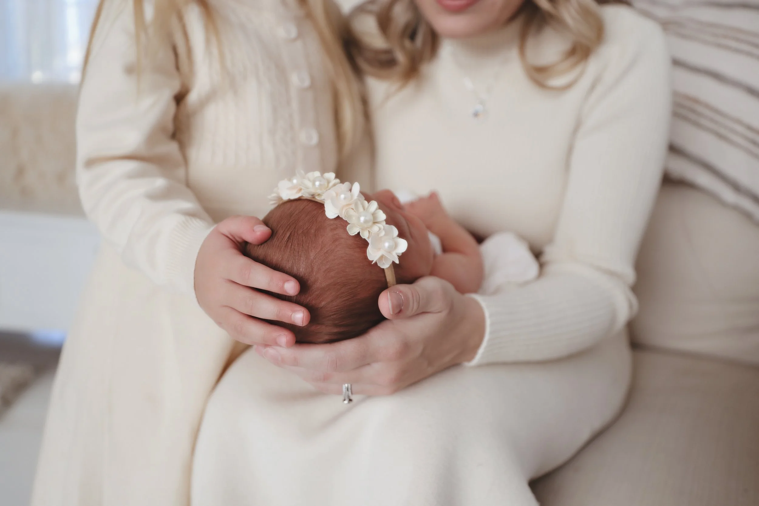 A woman holding a newborn baby girl with a floral headband in a cozy, cream-colored room.