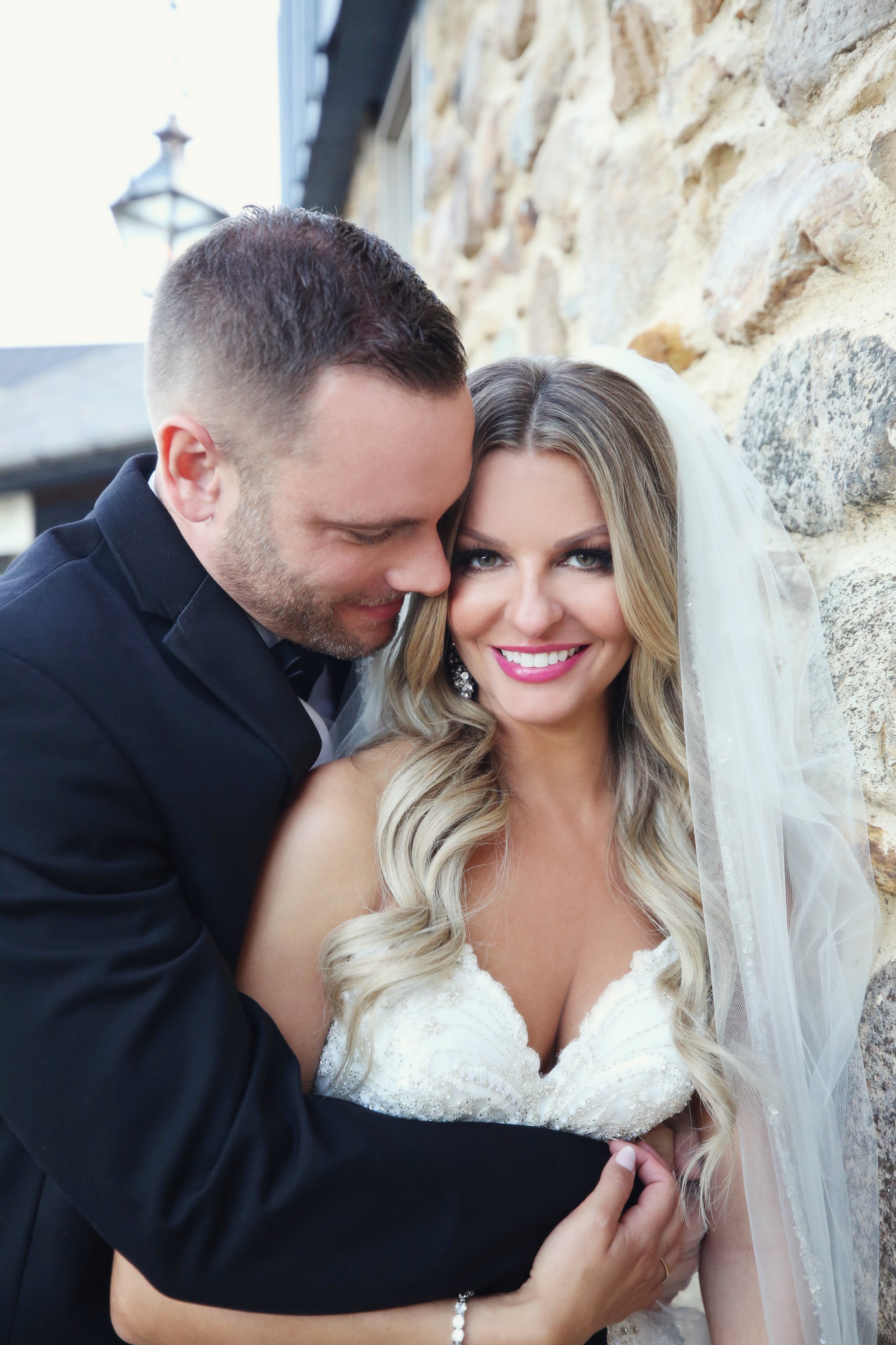 A newlywed couple in wedding attire posing outdoors. The groom is in a black suit and has his head close to the bride, who is smiling and wearing a white wedding dress with a veil, standing against a stone wall.