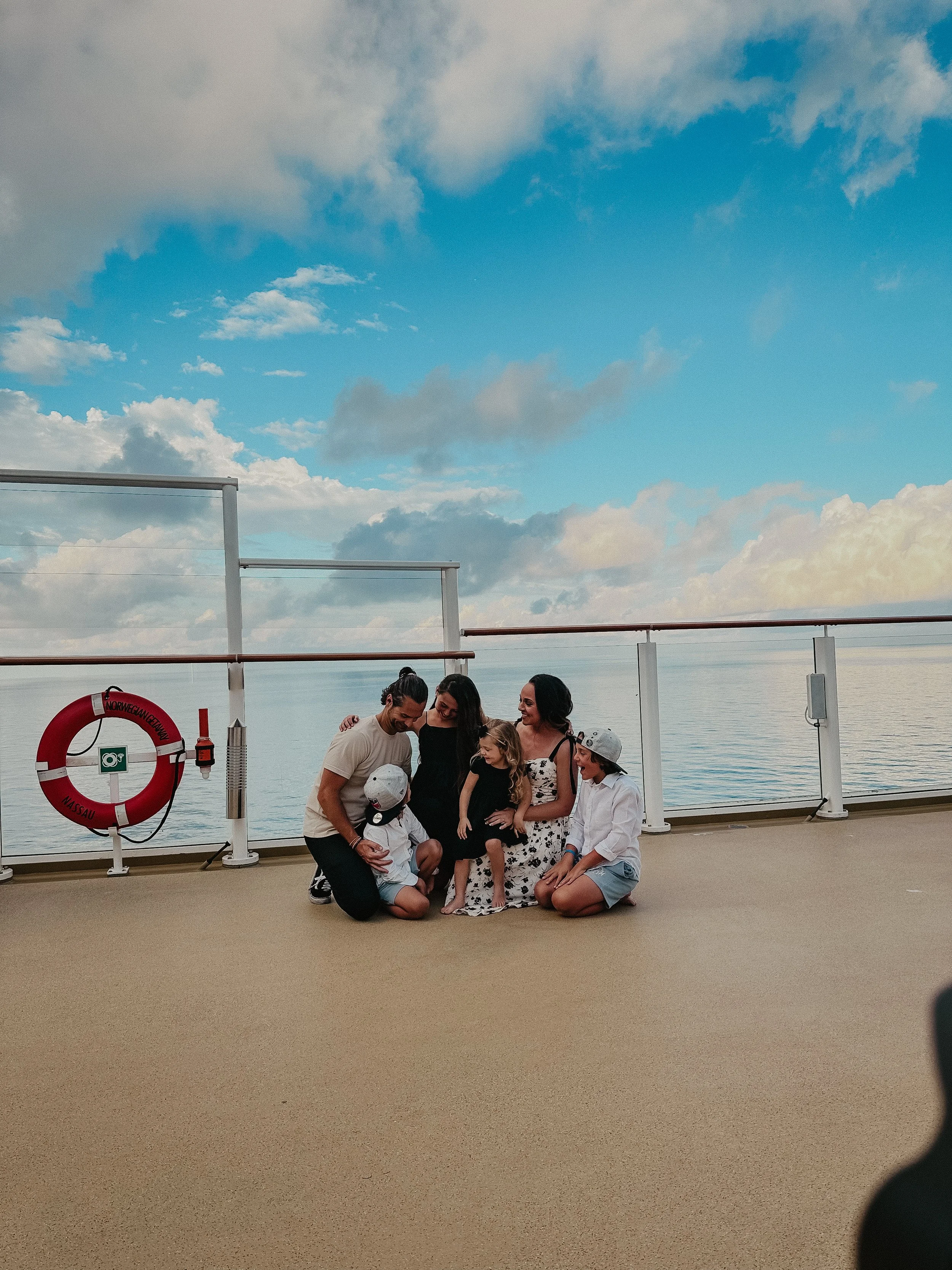 A group of six people, including children, sitting and kneeling on a deck by the water, with a blue sky and clouds in the background, enjoying a moment together.