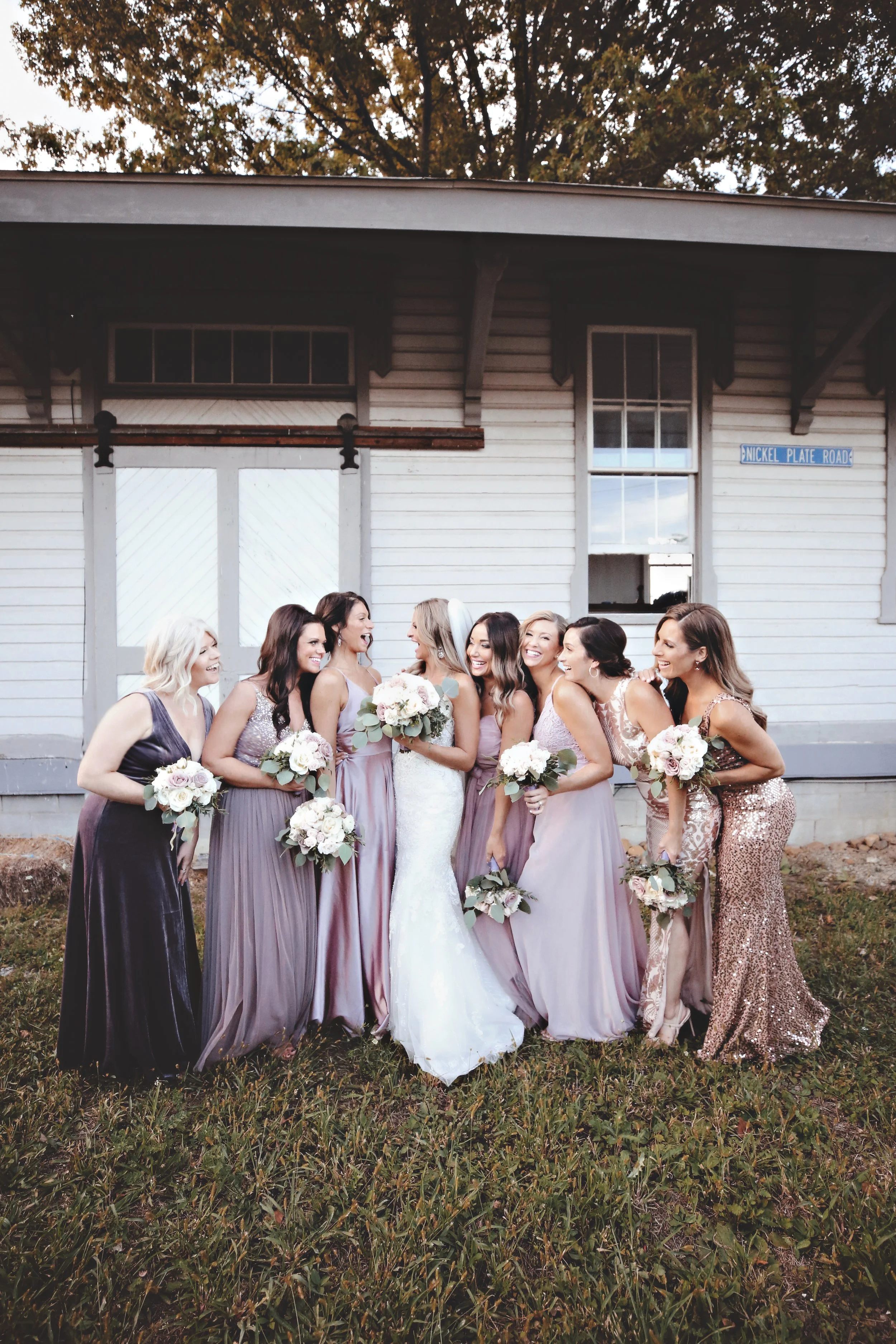 A bride and seven bridesmaids standing in front of a gray building with a sign that reads 'Nickel Plate Road,' holding bouquets of flowers and smiling at each other.