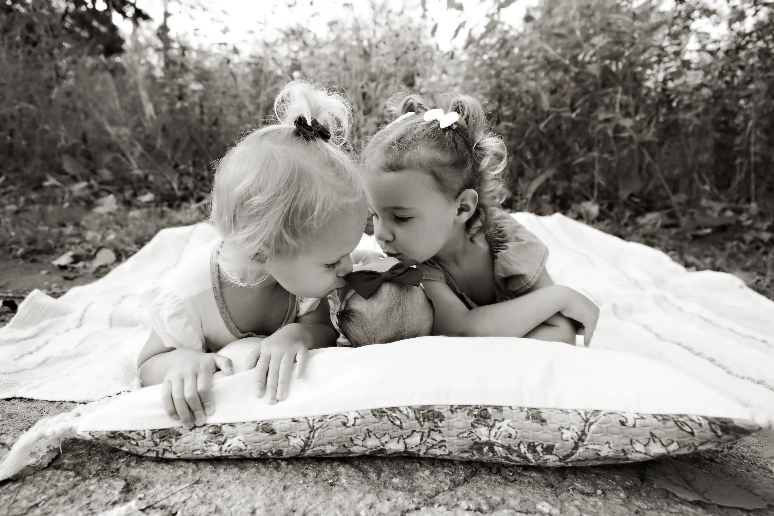 Three young children, two girls and a baby, lying on a blanket outdoors, nose to nose and gazing at each other, in a natural setting.