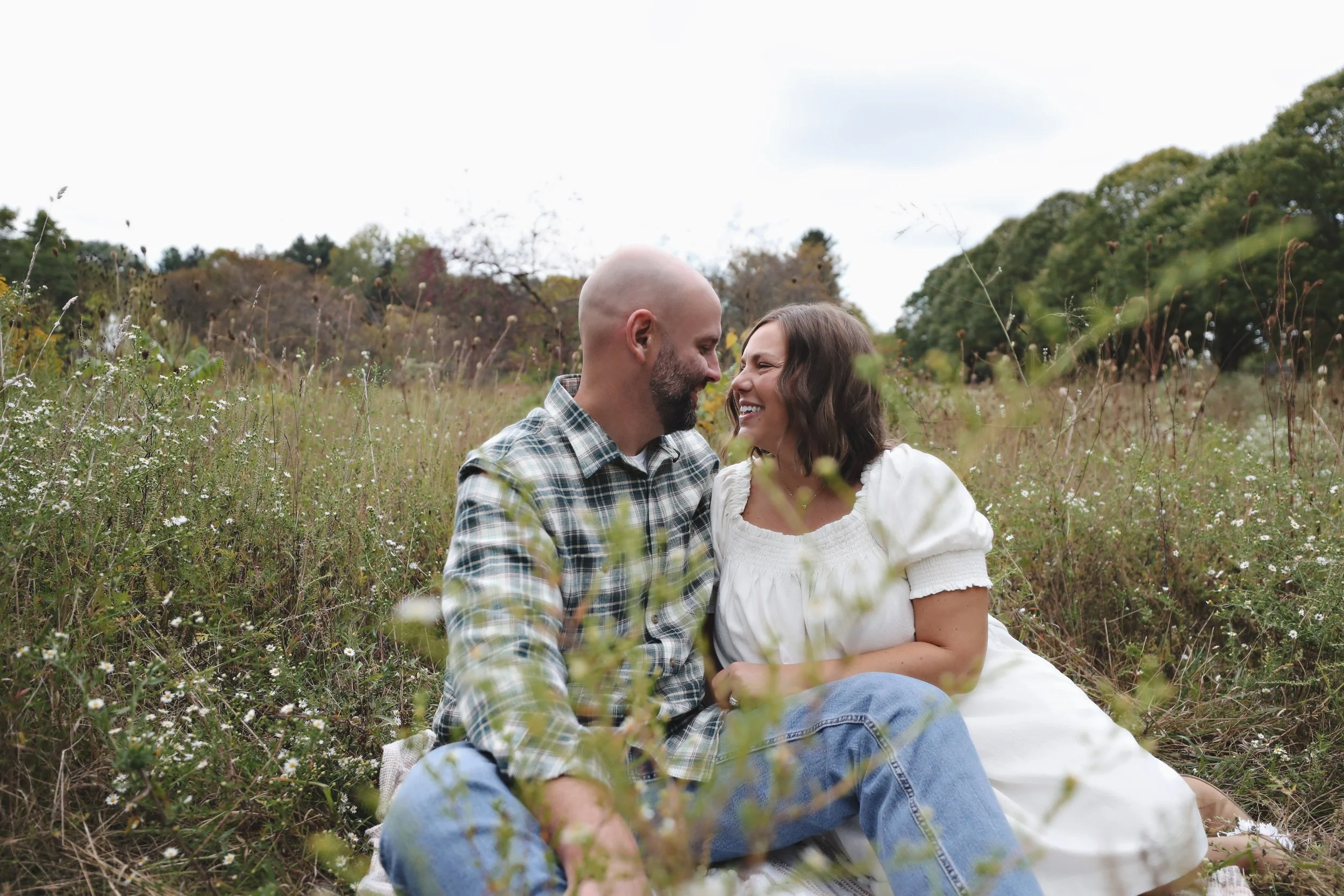 A couple sits in a grassy field with wildflowers, smiling and touching foreheads, surrounded by trees and a cloudy sky.