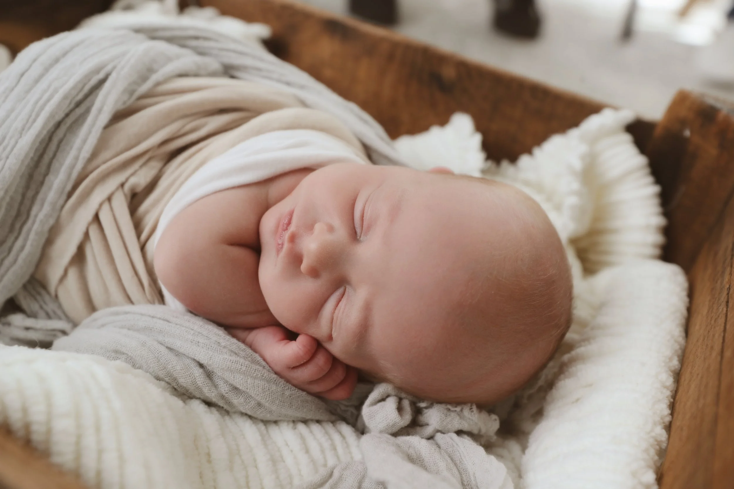 A sleeping baby wrapped in soft beige and white blankets, lying in a wooden cradle, with a cozy, relaxed expression.