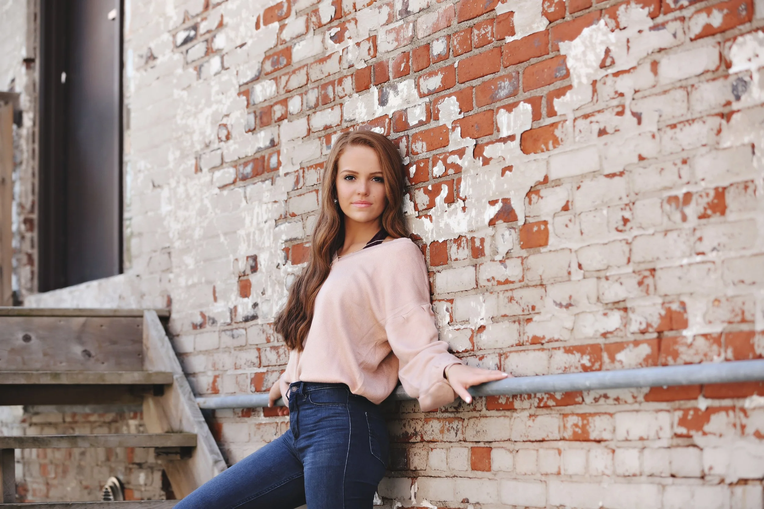 Young woman with long wavy brown hair in a pink blouse and blue jeans leaning against a weathered brick wall with metal railing.