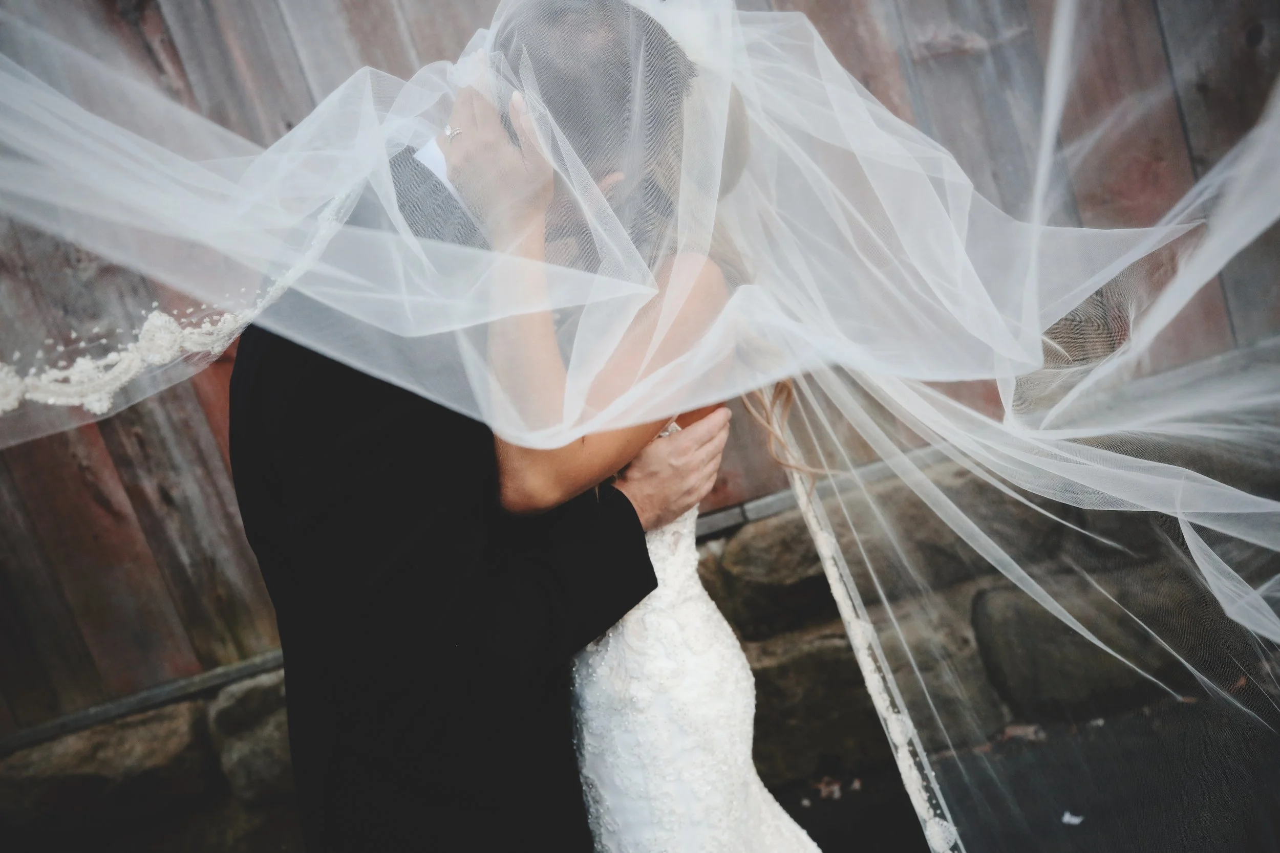 A couple sharing a kiss underneath a wedding veil, with the bride wearing a white lace wedding dress and the groom in a black suit, outdoors near a stone wall.