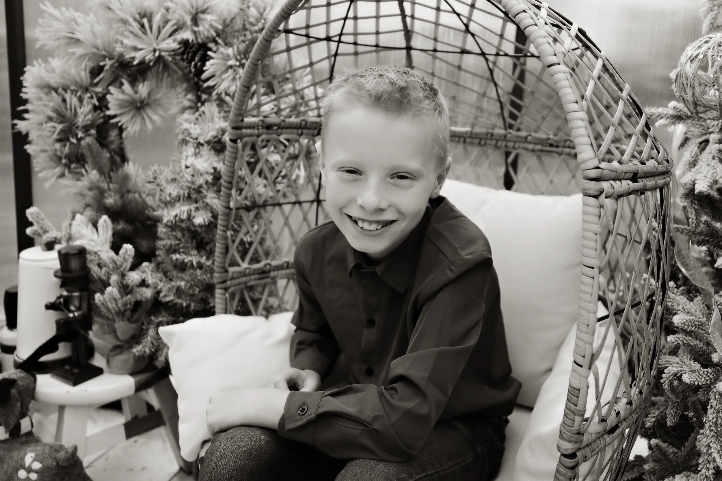 A young boy with a big smile sitting in a wicker chair surrounded by Christmas decorations, including a small Christmas tree and a wreath.