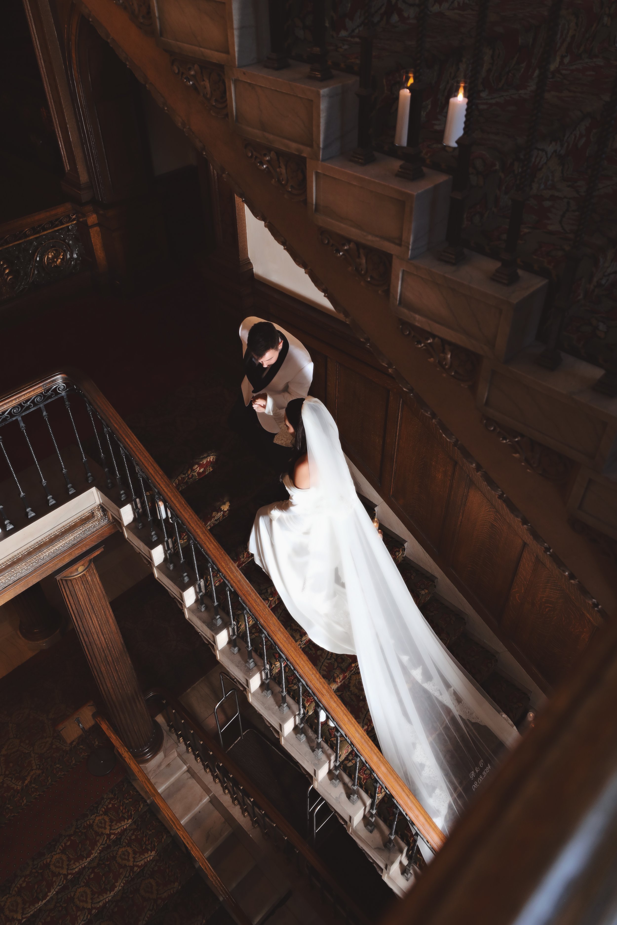 A bride and groom standing on a staircase looking at each other in a dimly lit, elegant indoor setting.