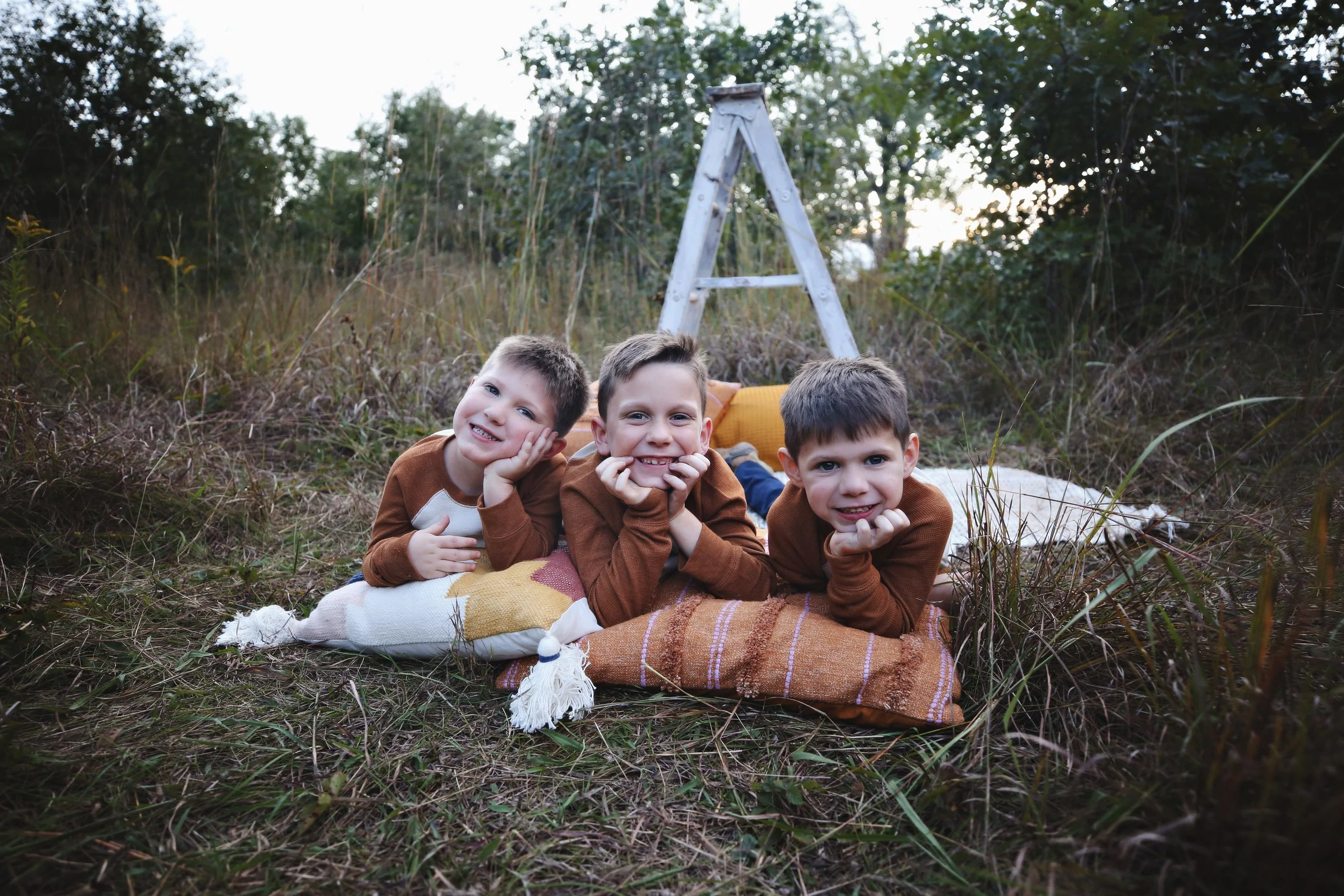 Three young boys lying on blankets outdoors in a grassy field, smiling and resting their chins on their hands, with a wooden ladder and trees in the background.