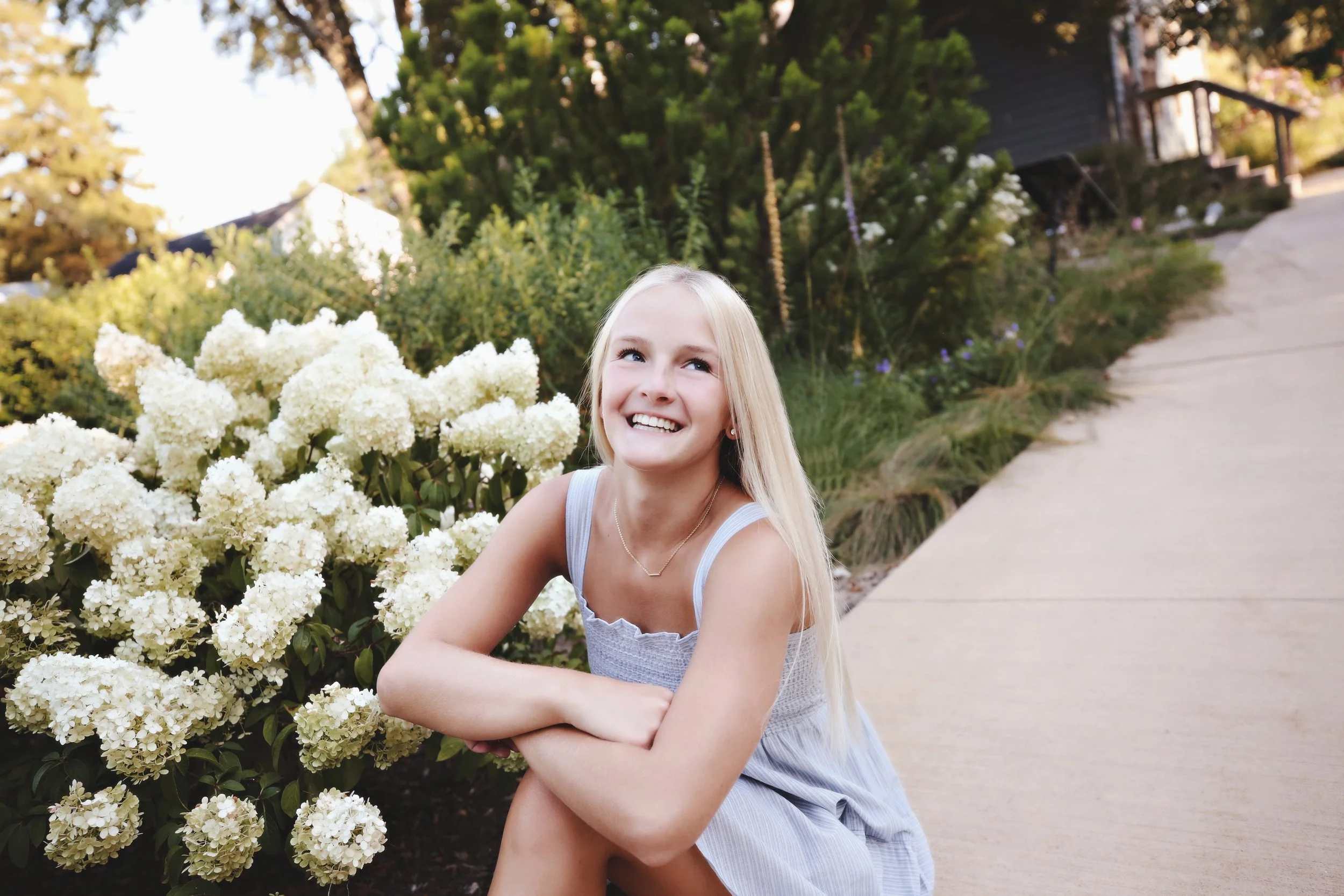 A young girl with long blonde hair smiling while sitting on a sidewalk next to white hydrangea flowers in a garden.