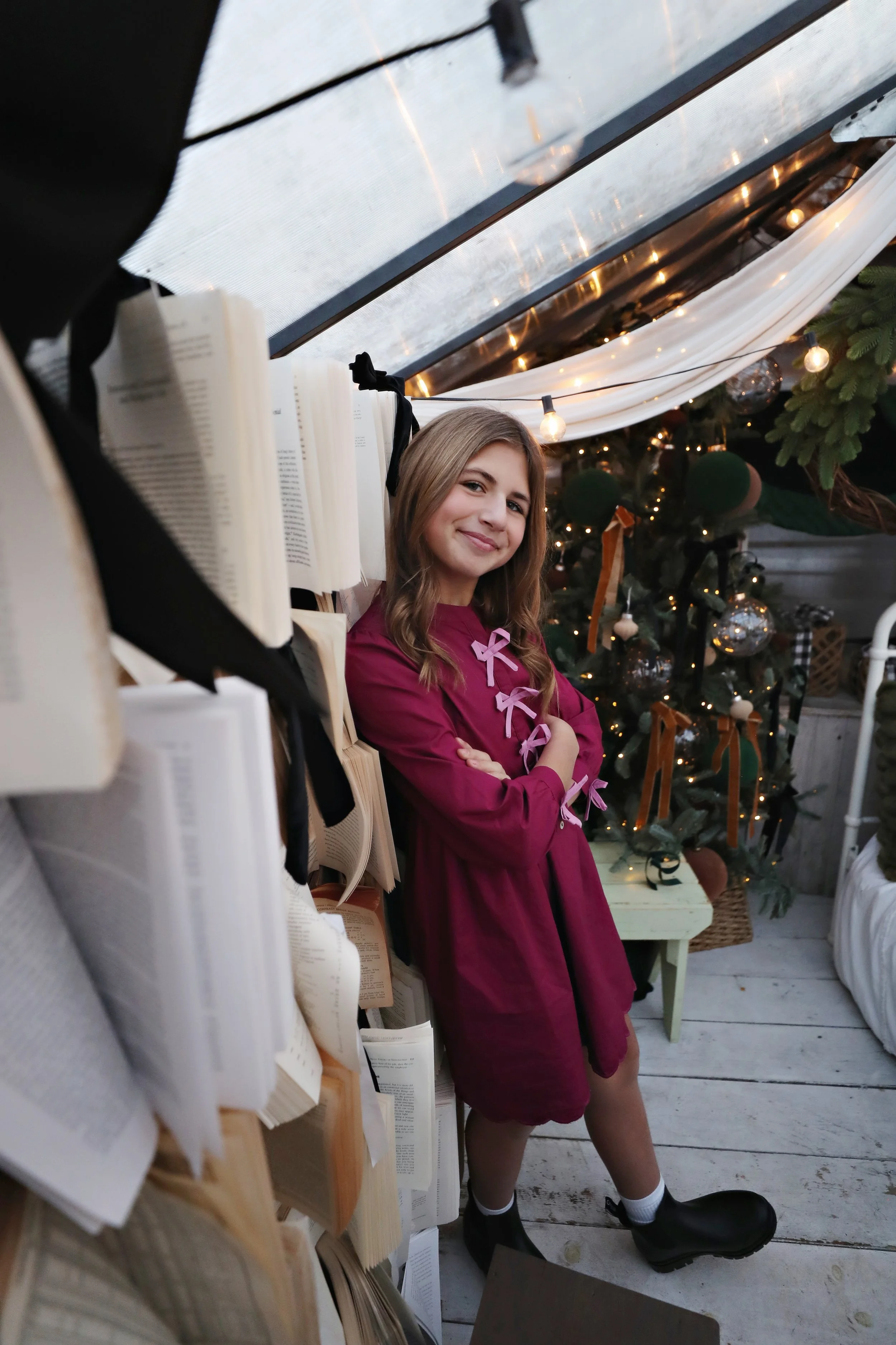 A young woman with wavy light brown hair and a warm smile, wearing a magenta dress with pink bows, standing next to a bookshelf filled with open books, in a cozy, decorated indoor space with a Christmas tree in the background adorned with lights, orn