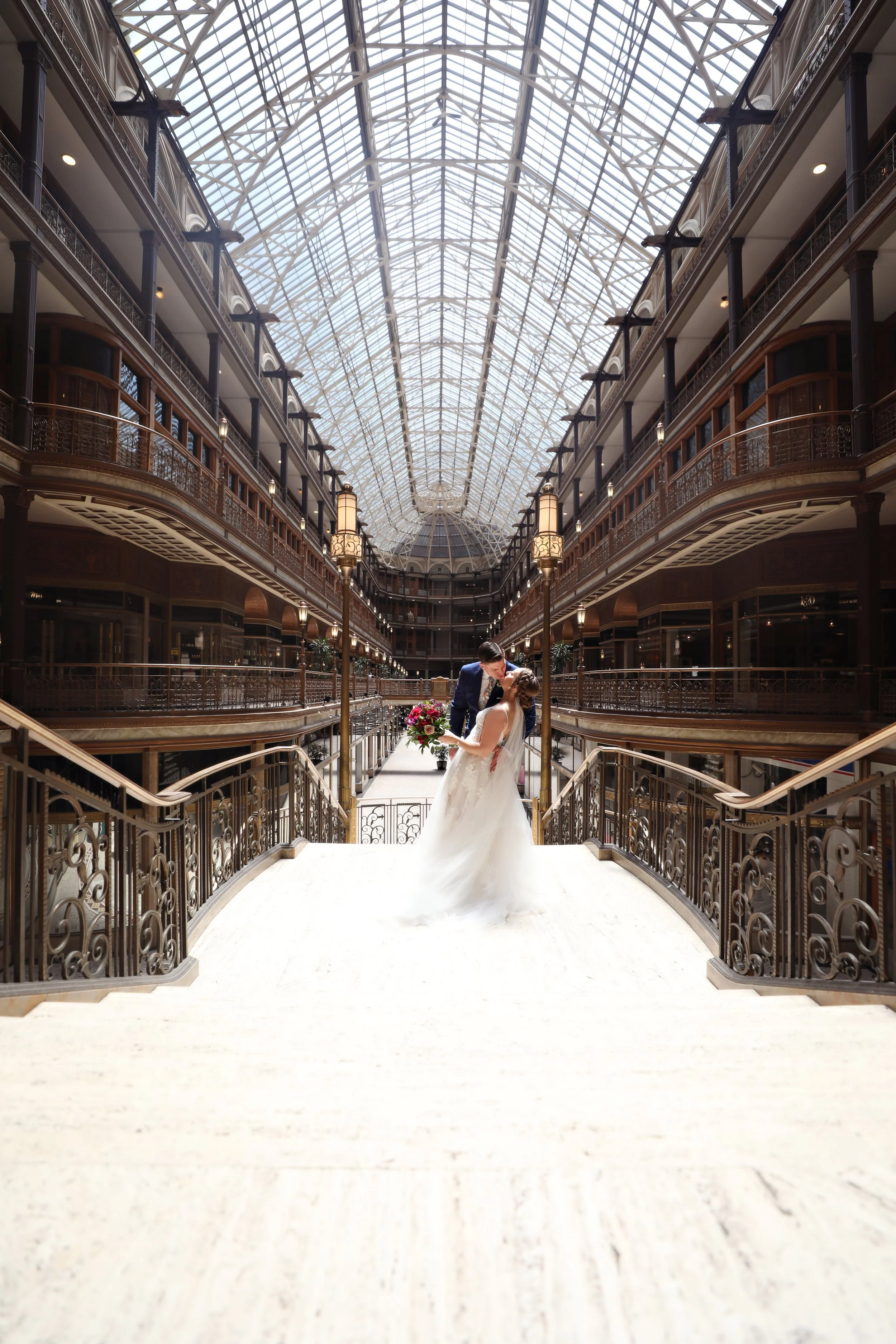 A bride and groom sharing a kiss inside a grand glass-ceilinged shopping mall or hotel atrium, with the bride holding a bouquet of flowers.