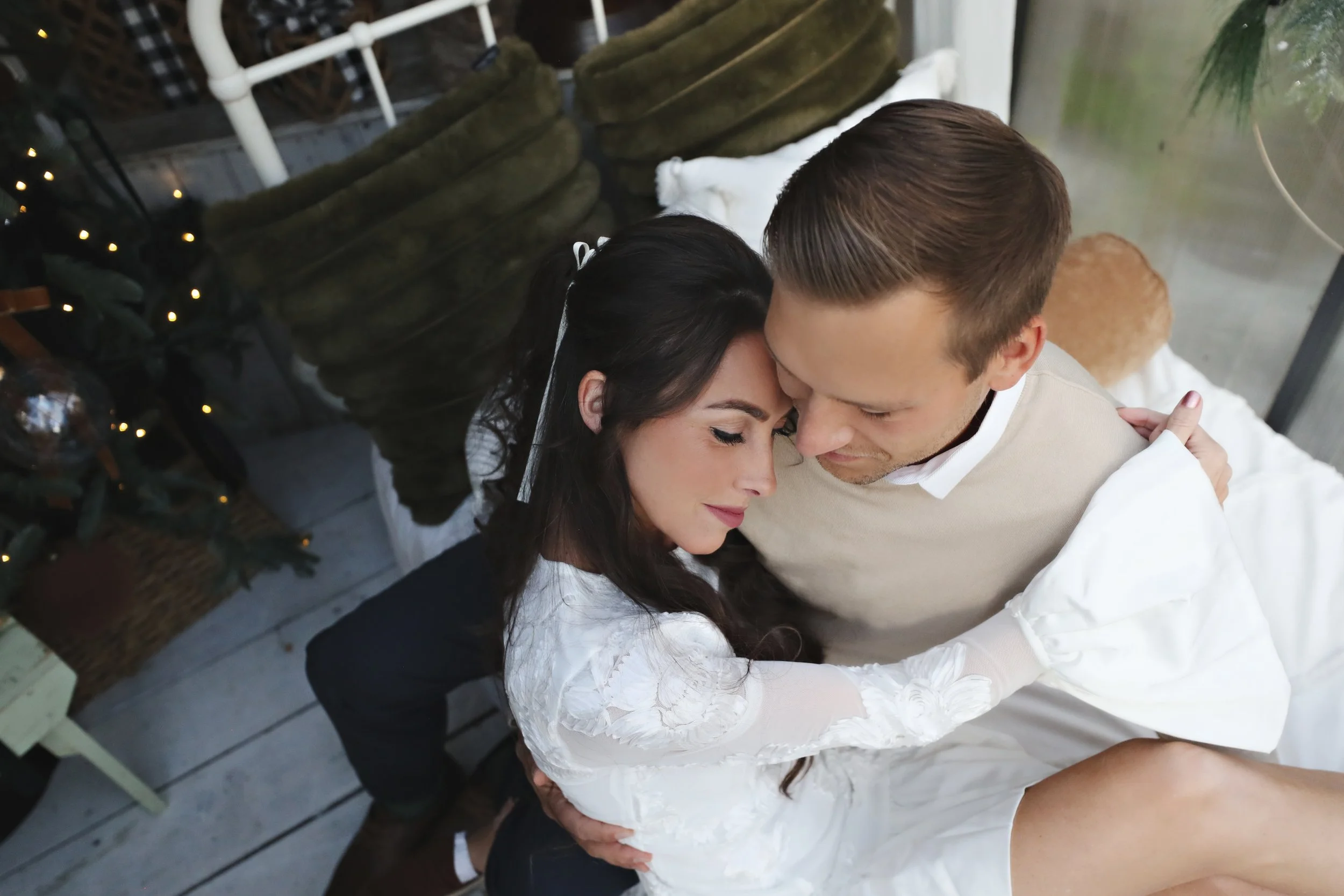 A couple in a warm embrace, sitting closely with their foreheads touching, in a cozy, decorated indoor space.