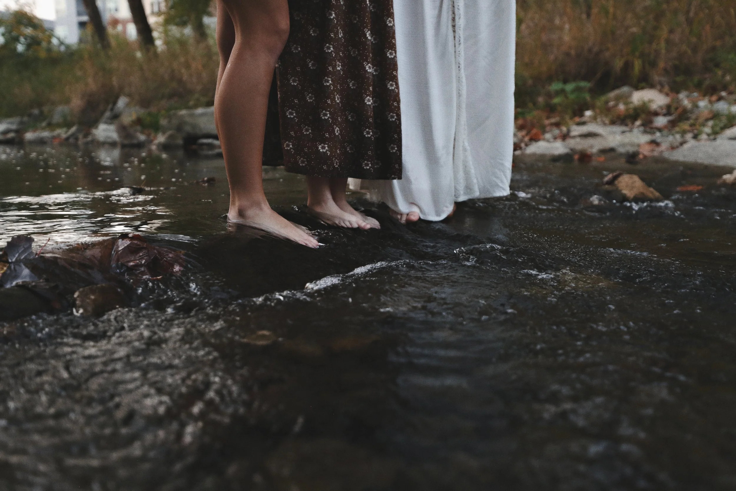 Two people standing in a shallow creek with bare feet, surrounded by rocks and autumn-colored trees.