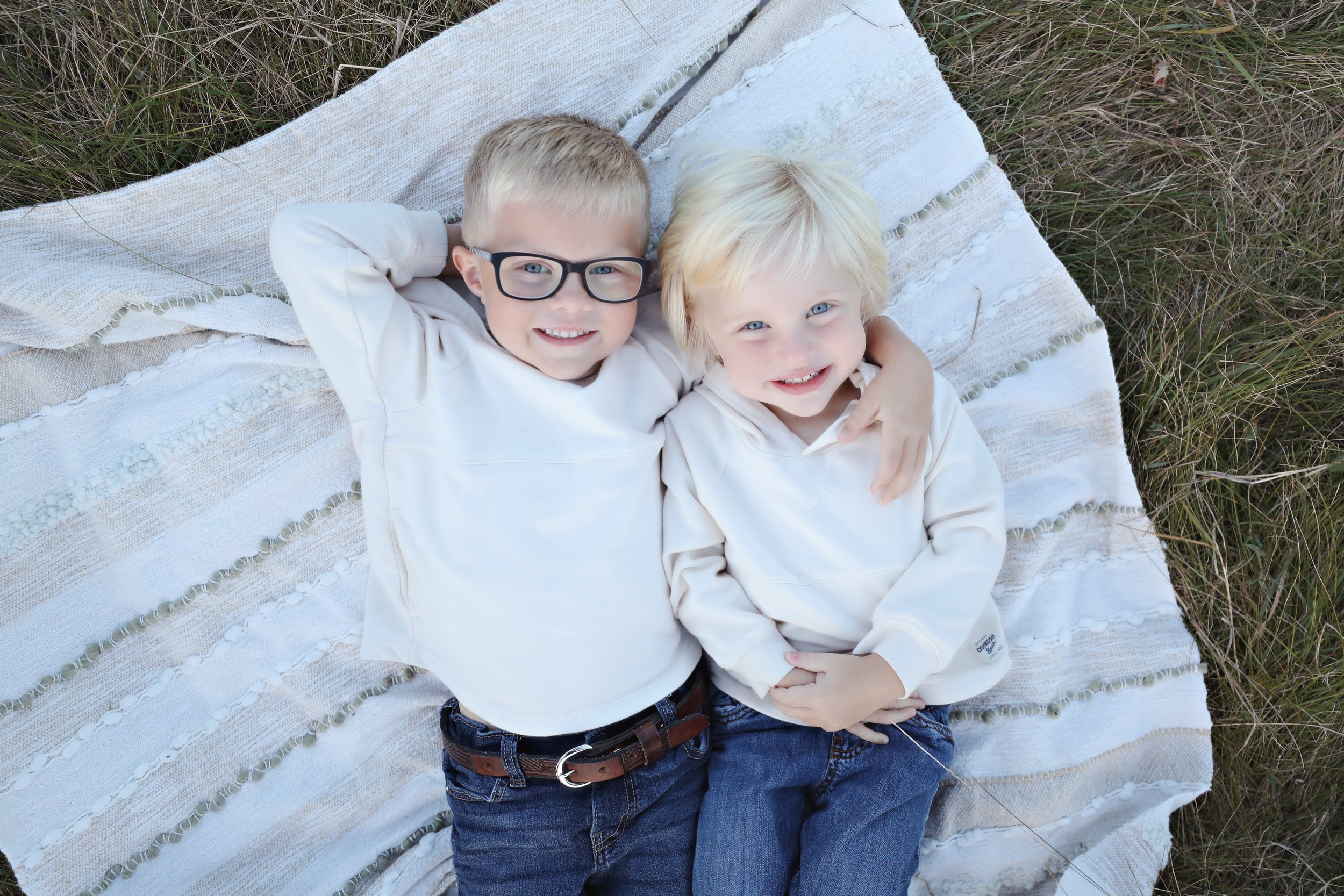 Two young boys with blonde hair and blue eyes lying on a blanket outdoors, smiling and looking up at the camera. One boy wears glasses and a white sweatshirt, and the other has a white hoodie.