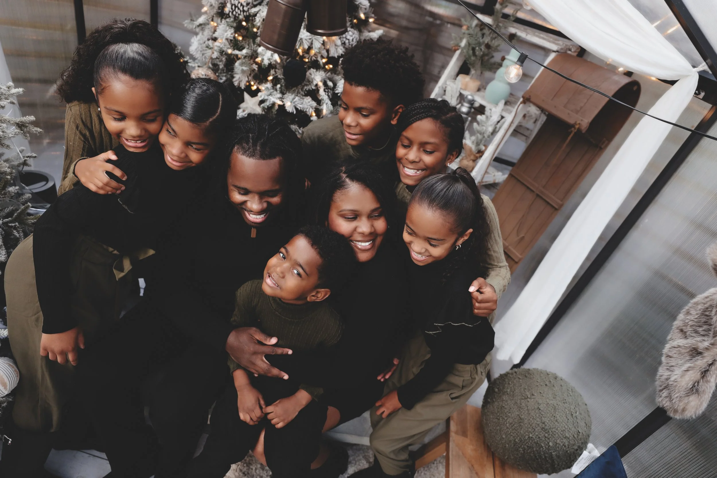 A group of nine people, including children and adults, gathered closely together indoors in front of a decorated Christmas tree, smiling and enjoying each other's company during the holiday season.