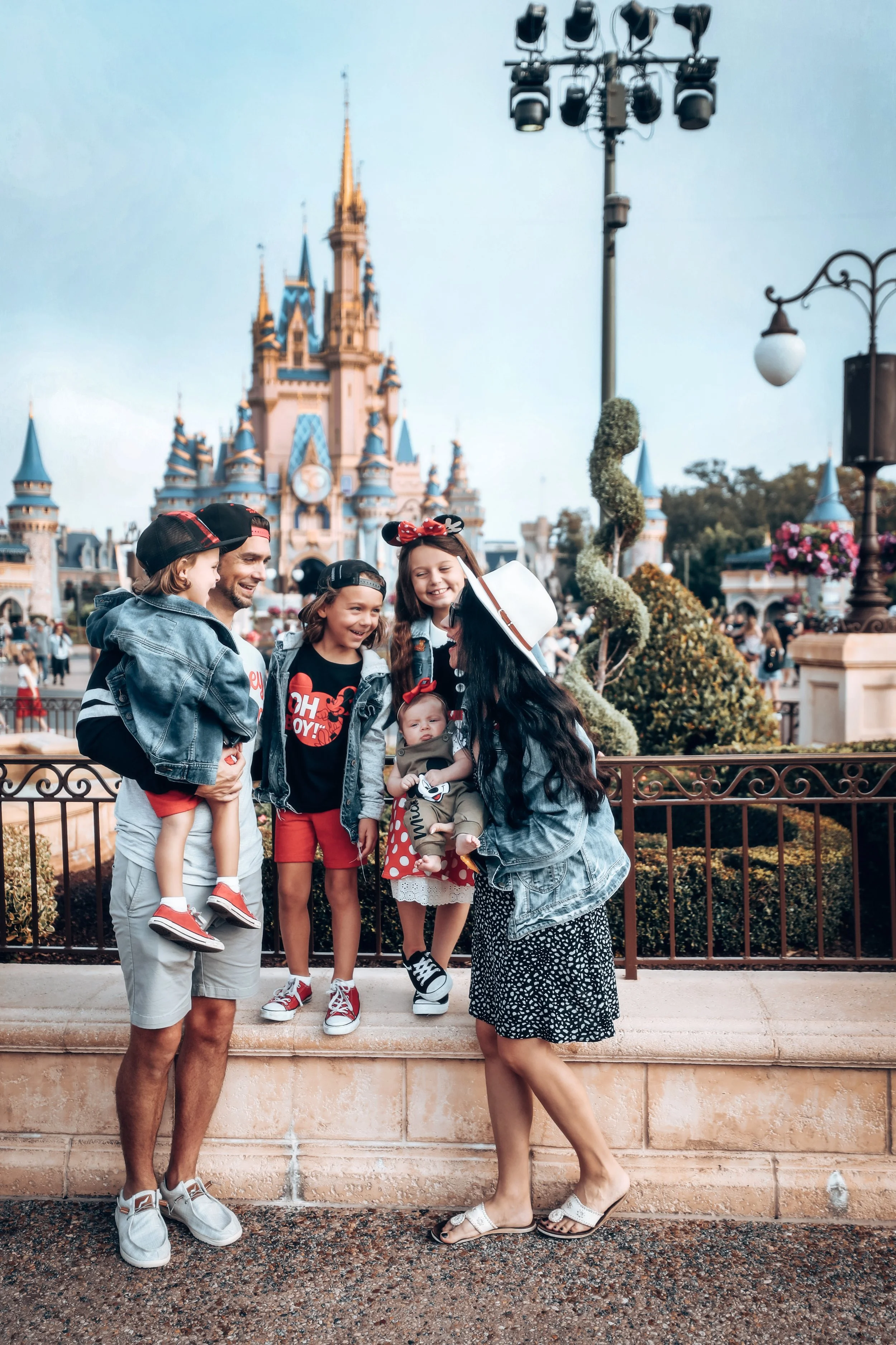 Smiling family of five standing in front of Sleeping Beauty Castle at Disneyland, California, posing for a photo together, with trees, decorative topiary, and park visitors in the background.