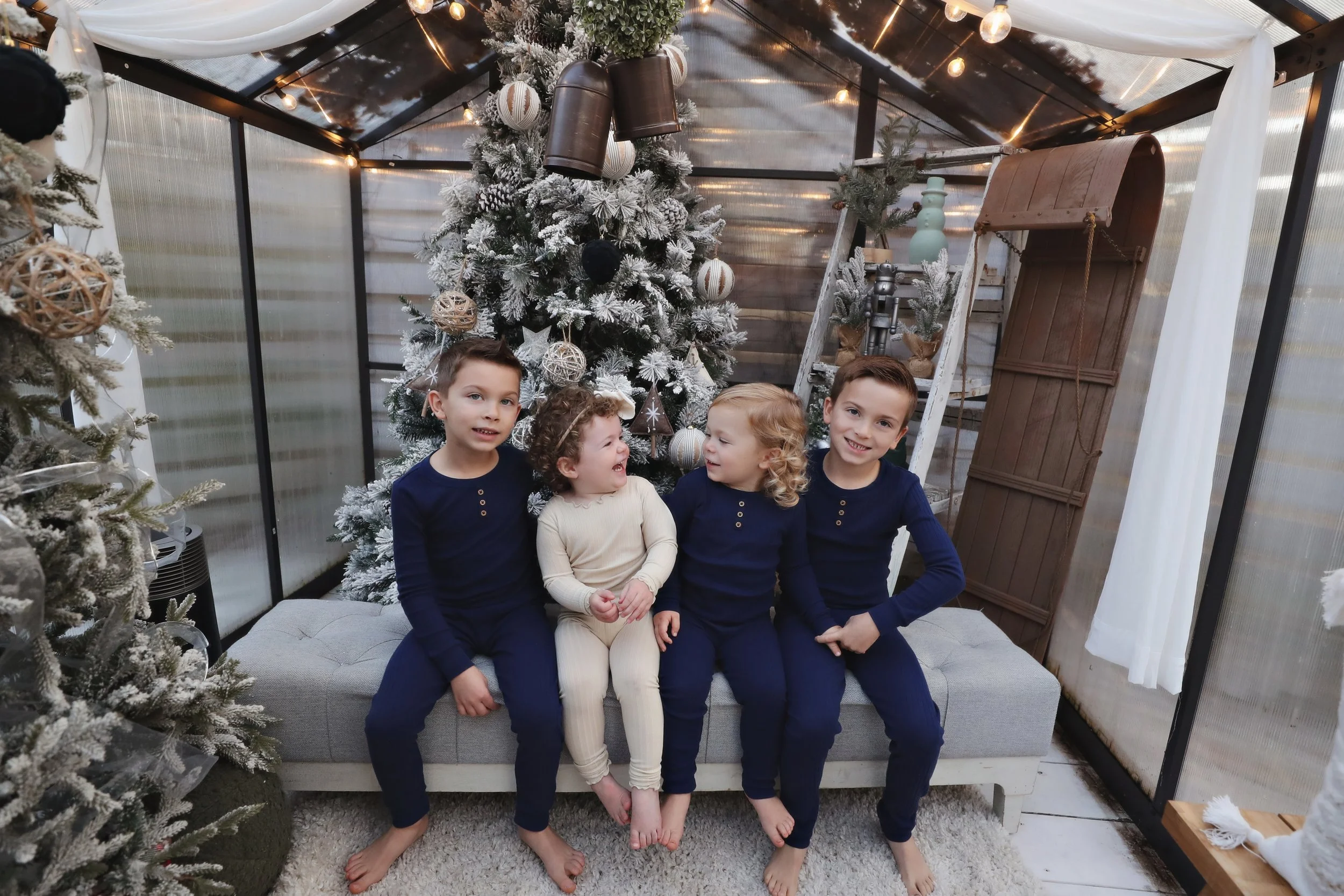 Four children sitting on a grey sofa in front of a decorated Christmas tree inside a greenhouse-shaped room. The children are smiling and wearing navy and beige pajamas, with a snow-flocked Christmas tree, ornaments, and warm lighting in the backgrou